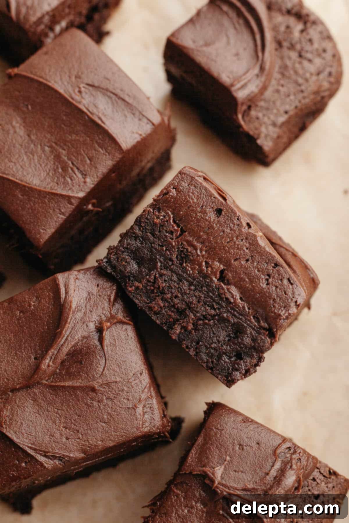 Rich Chocolate Brownies with Tangy Cream Cheese Frosting 2 Brownies covered in chocolate cream cheese frosting on parchment paper, ready to be sliced and enjoyed.