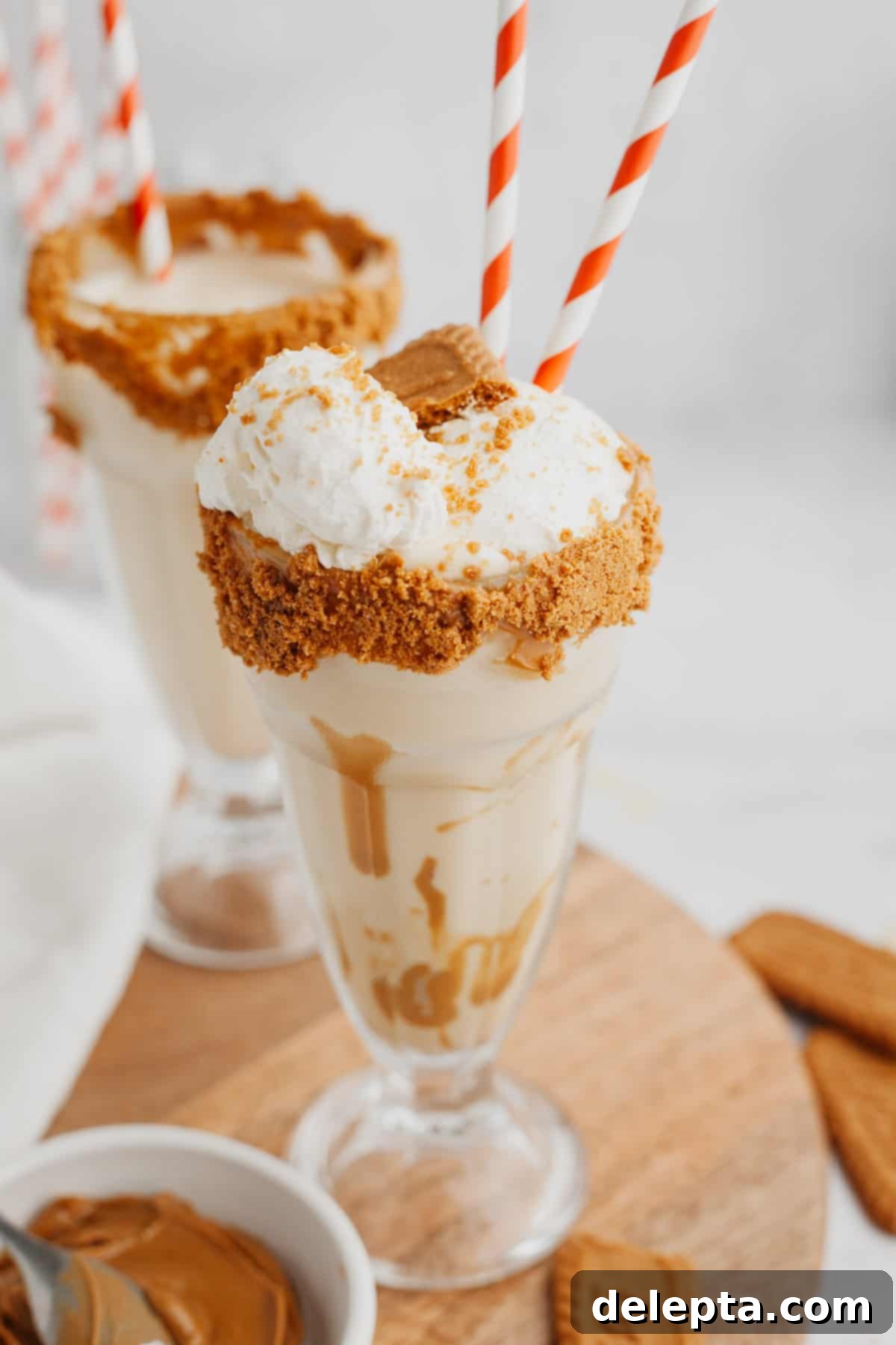Creamy Biscoff Milkshake 2 Two biscoff milkshakes in large glasses with red and white straws, topped with whipped cream and crushed cookies.