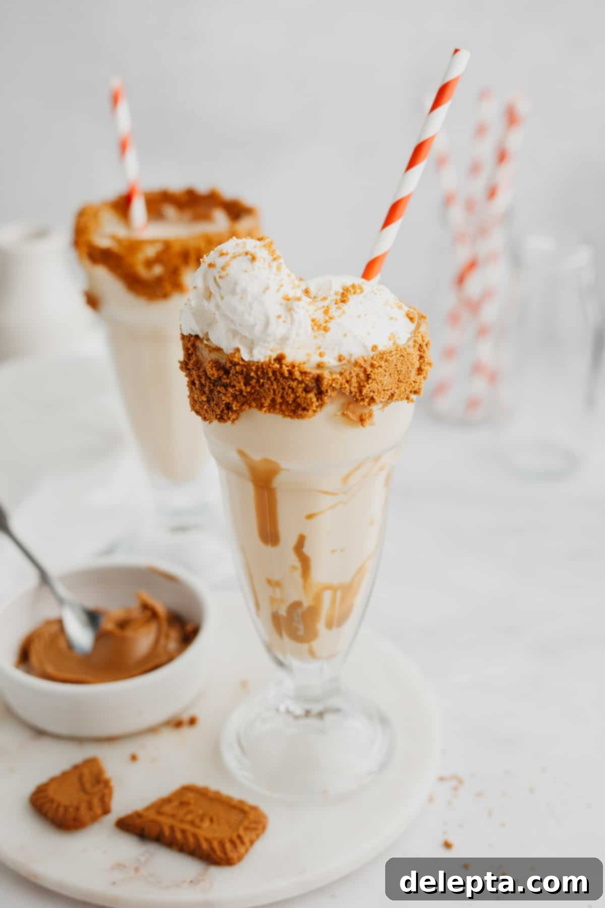 Creamy Biscoff Milkshake 5 Two biscoff milkshakes in large glasses with red and white straws, garnished with whipped cream and crushed biscoff cookies.