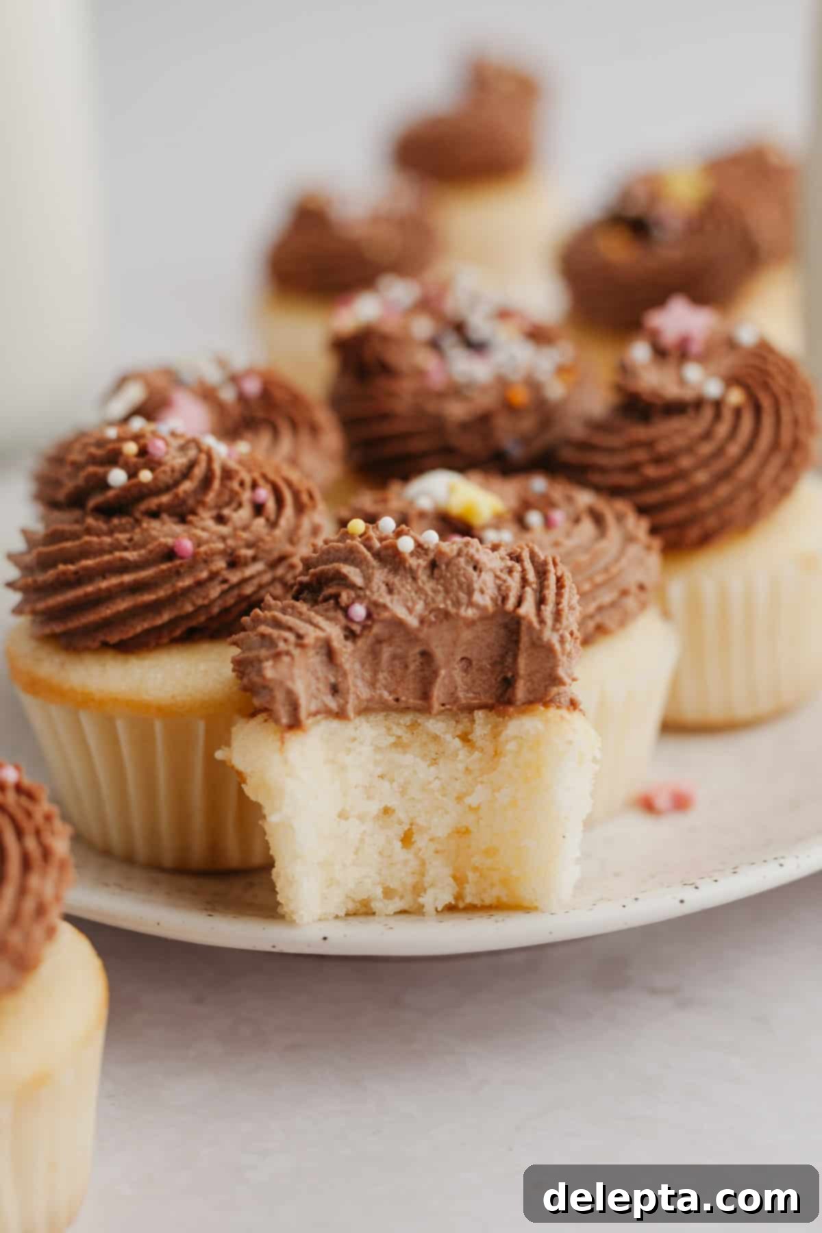 Dainty Vanilla Delights 7 A close-up shot of a single mini vanilla cupcake, elegantly piped with chocolate frosting. A bite has been taken, revealing the moist, light vanilla cake underneath.