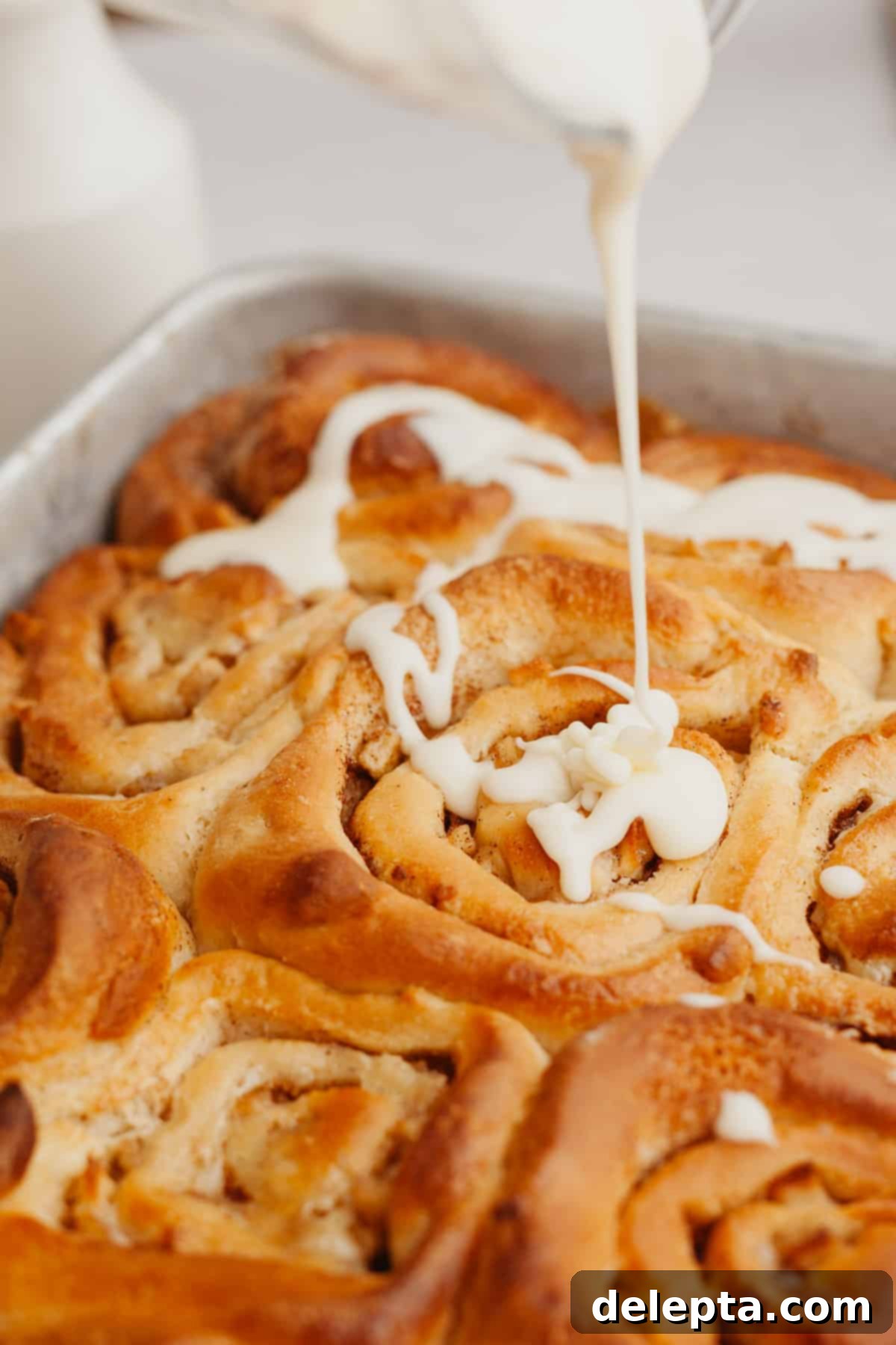 Apple Pie Stuffed Cinnamon Rolls 9 Maple icing being drizzled generously onto warm cinnamon rolls in a baking pan.