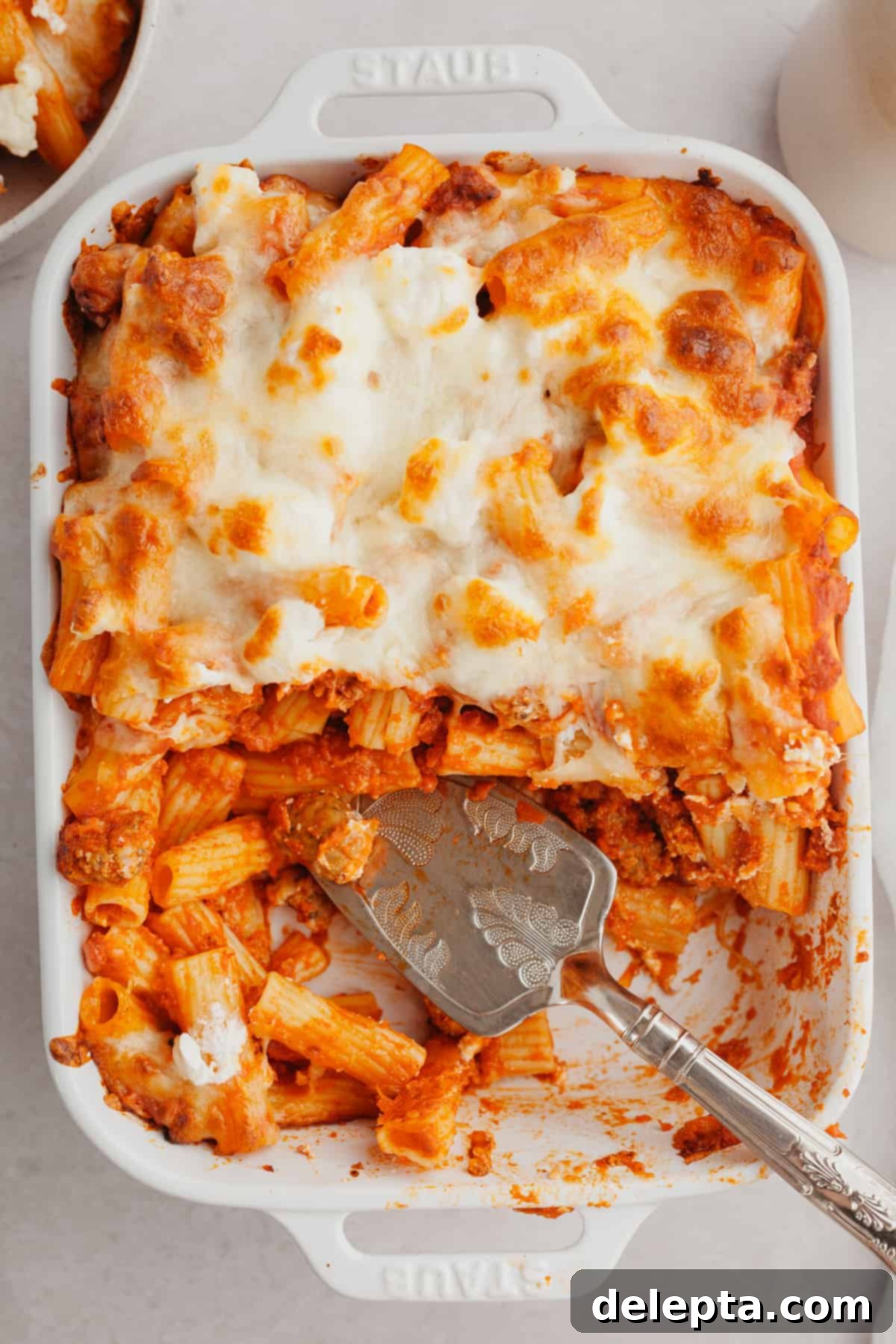 Hearty Three-Cheese Baked Rigatoni with Savory Sausage 7 A white baking dish with a cheesy baked pasta in it, with a silver serving knife ready for serving.