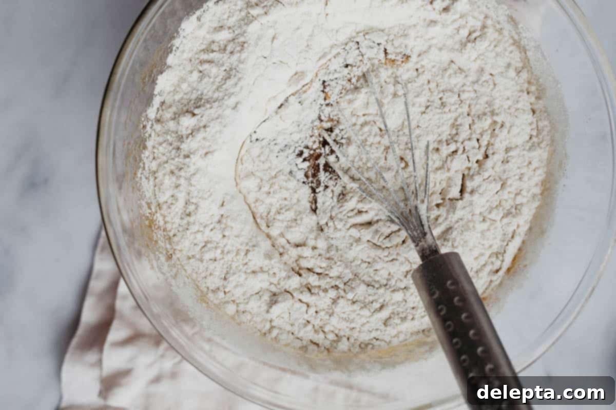 Sprinkle-Studded Golden Oreo Gooey Bars 6 Overhead view of flour and other dry ingredients being poured into the wet blondie batter in a mixing bowl, next to a whisk.