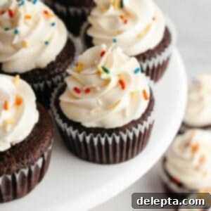 Velvet Chocolate Cupcakes with Tangy Cream Cheese Swirl 12 Chocolate cupcakes with a white frosting and rainbow sprinkles on a cake stand.