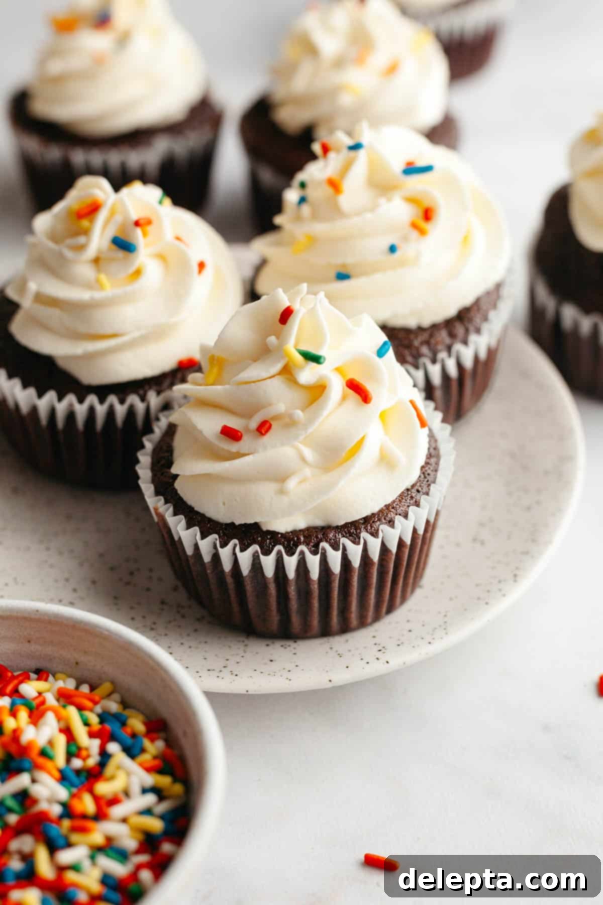 Velvet Chocolate Cupcakes with Tangy Cream Cheese Swirl 10 Three perfectly frosted chocolate cupcakes with white cream cheese frosting and rainbow sprinkles on a small white plate.