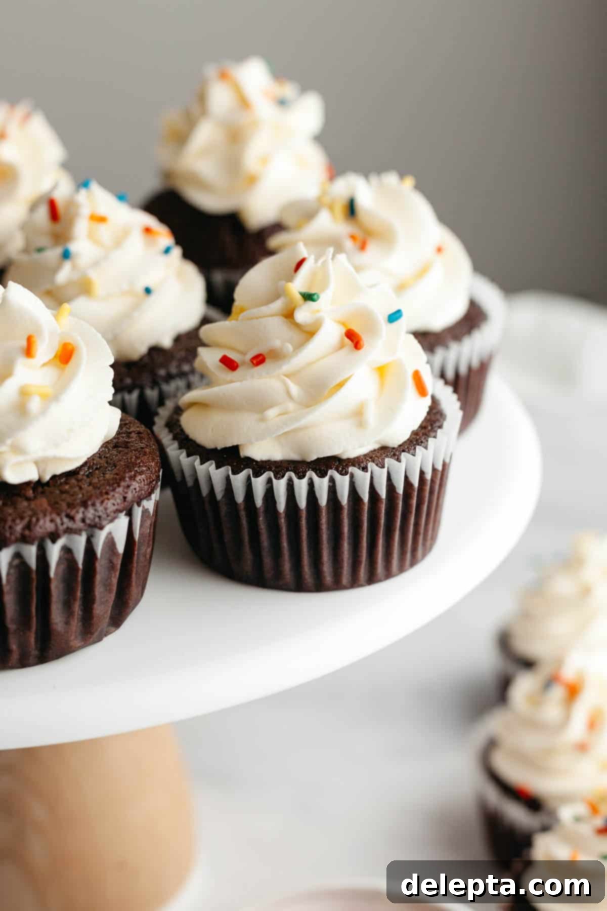 Perfect Butter-Free Cream Cheese Frosting in Minutes 7 Chocolate cupcakes with a white frosting and rainbow sprinkles on a cake stand, ready to be served.