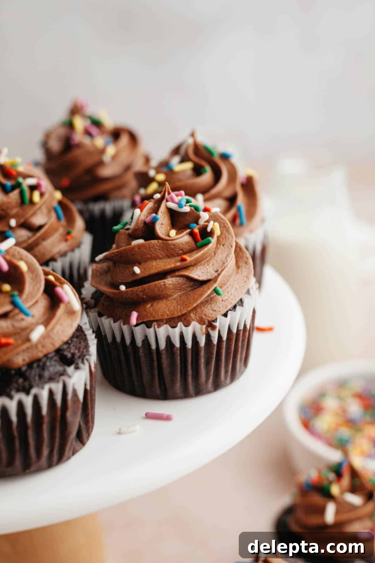 Decadent Chocolate Cream Cheese Cupcakes 11 Chocolate cupcakes with chocolate frosting and rainbow sprinkles artfully arranged on a white cake stand, ready for serving.