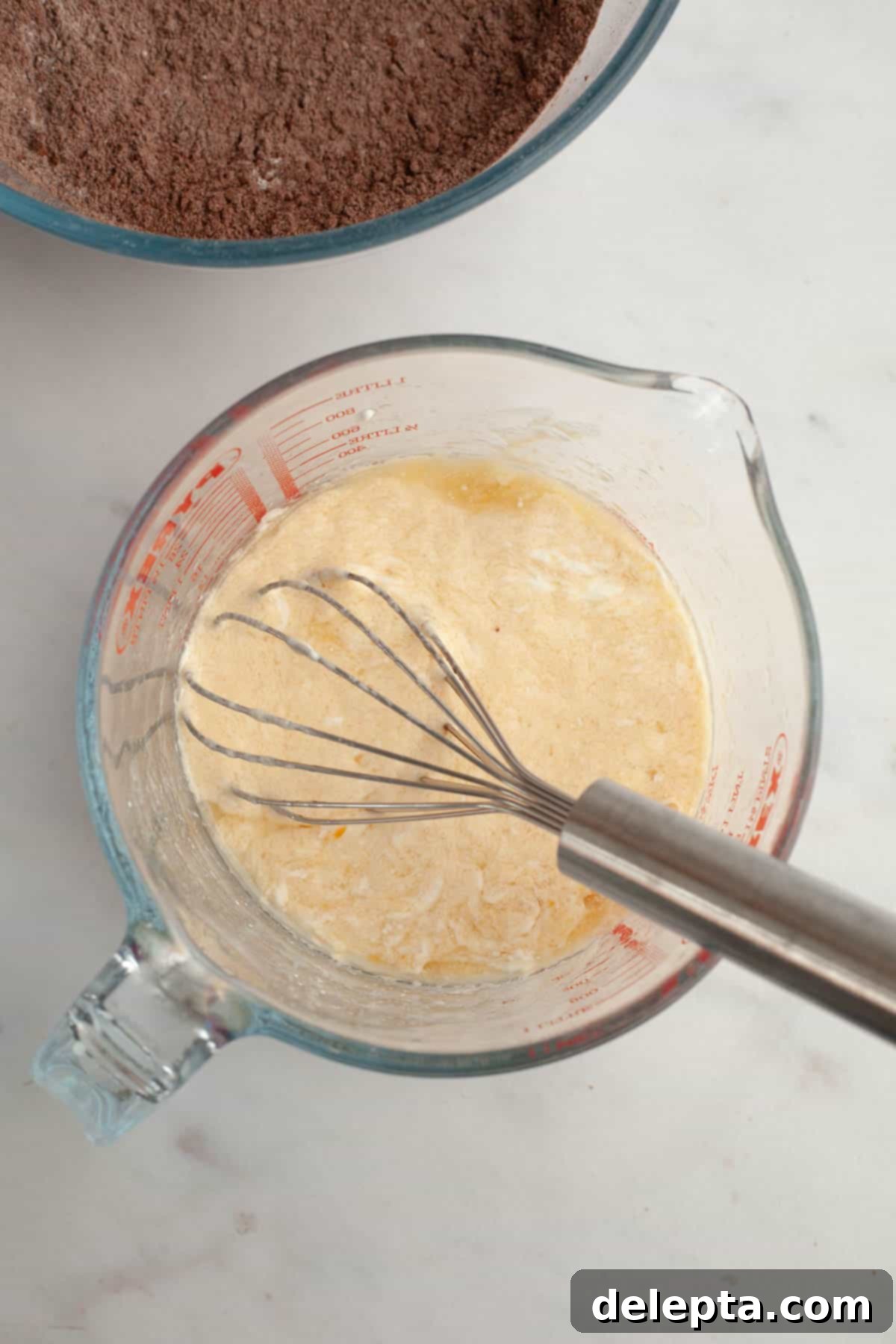 Decadent Chocolate Cream Cheese Cupcakes 5 A glass jug with a silver whisk and a light yellow mixture in it, representing wet ingredients.