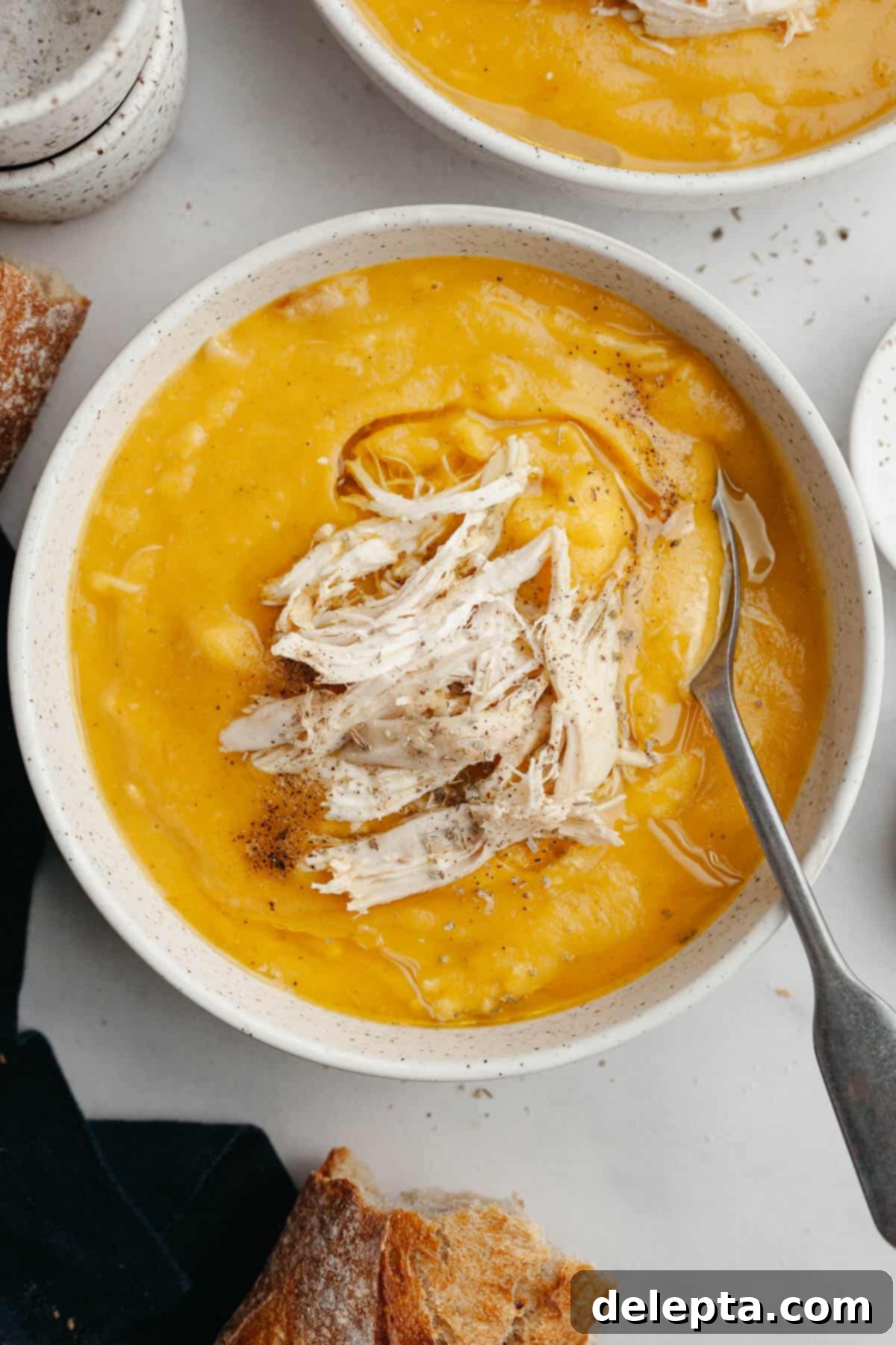 Autumn Gold Roasted Butternut Chicken Soup 11 A white speckled bowl filled with butternut squash chicken soup, accented with a silver spoon and ready to be enjoyed.