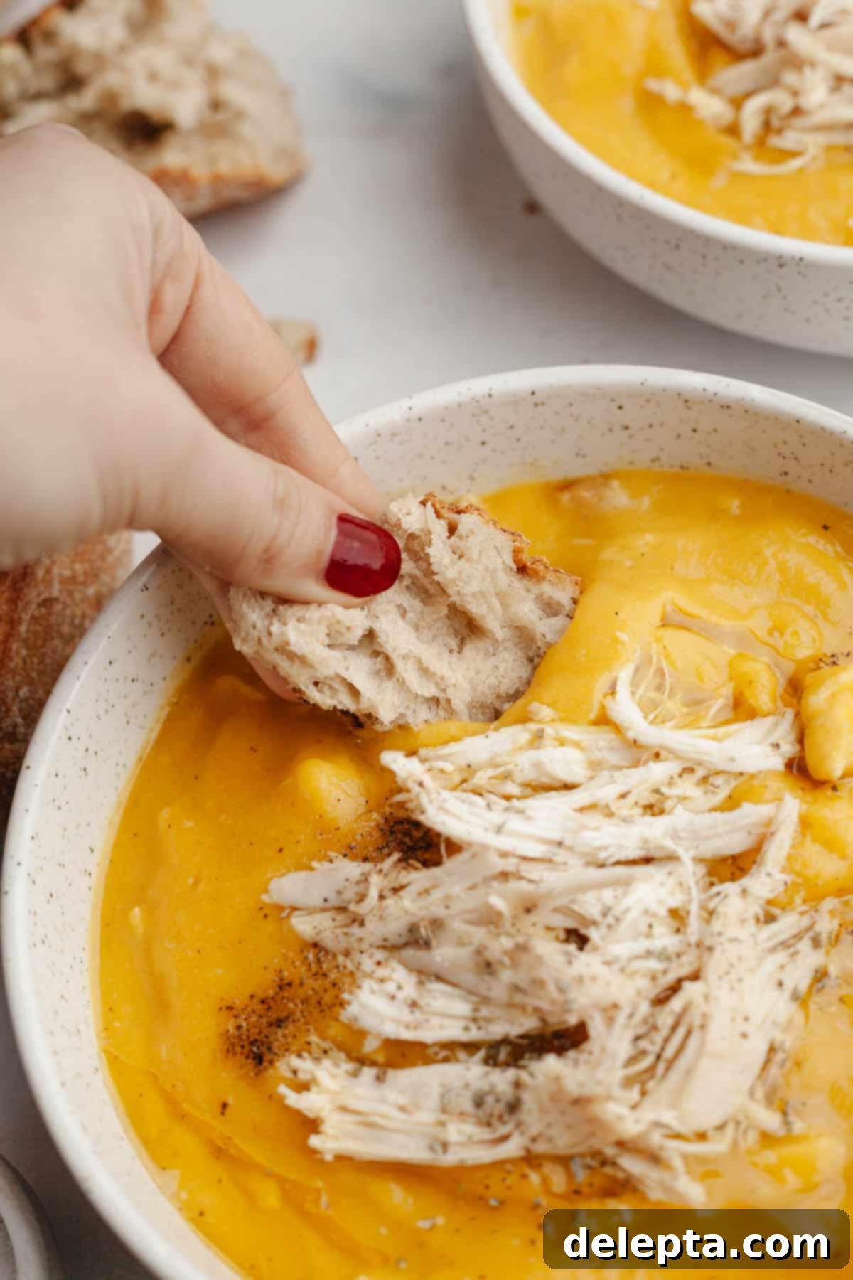 Autumn Gold Roasted Butternut Chicken Soup 10 A white bowl of butternut squash chicken soup with a hand dipping a piece of crusty bread into it, highlighting its comforting nature.
