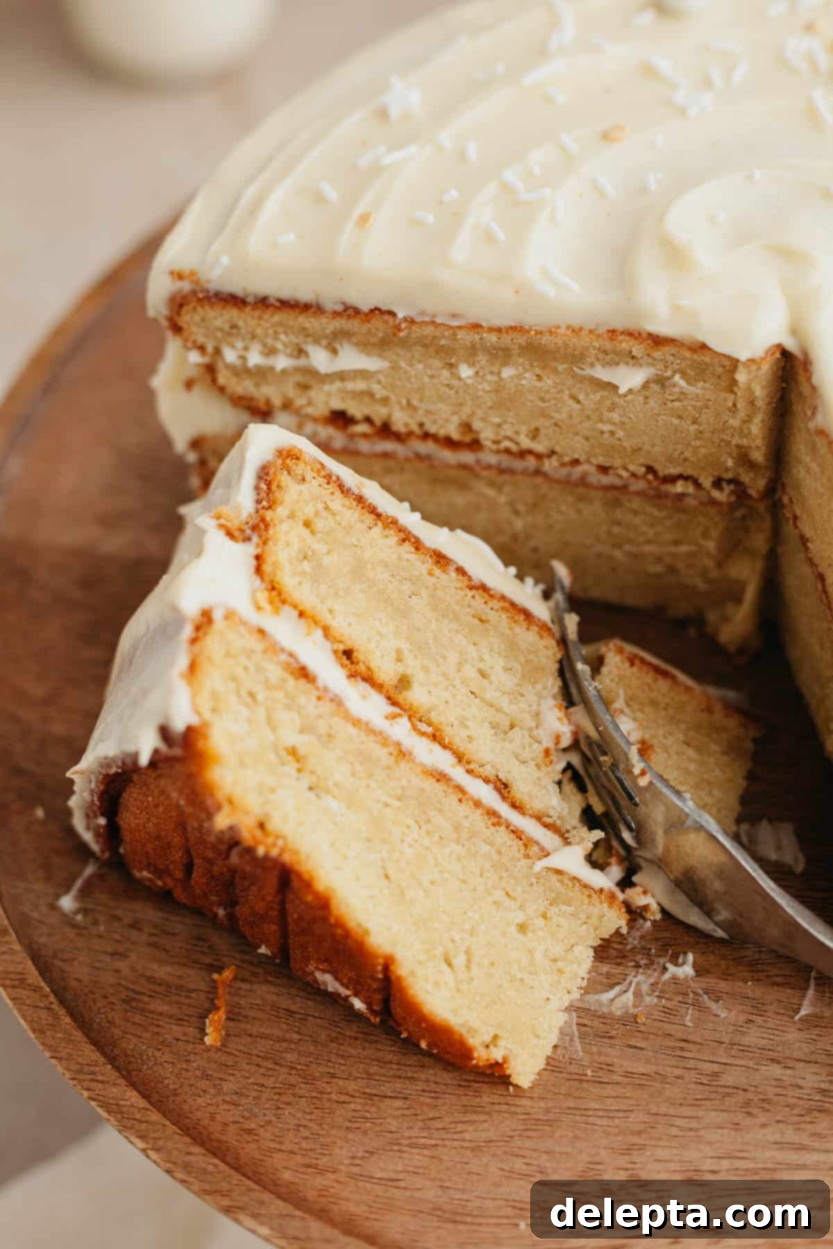 Brown Butter Cream Cheese Cardamom Cake 10 A beautifully decorated layer cake with white frosting on a rustic wooden cake stand, with one slice elegantly placed on its side.