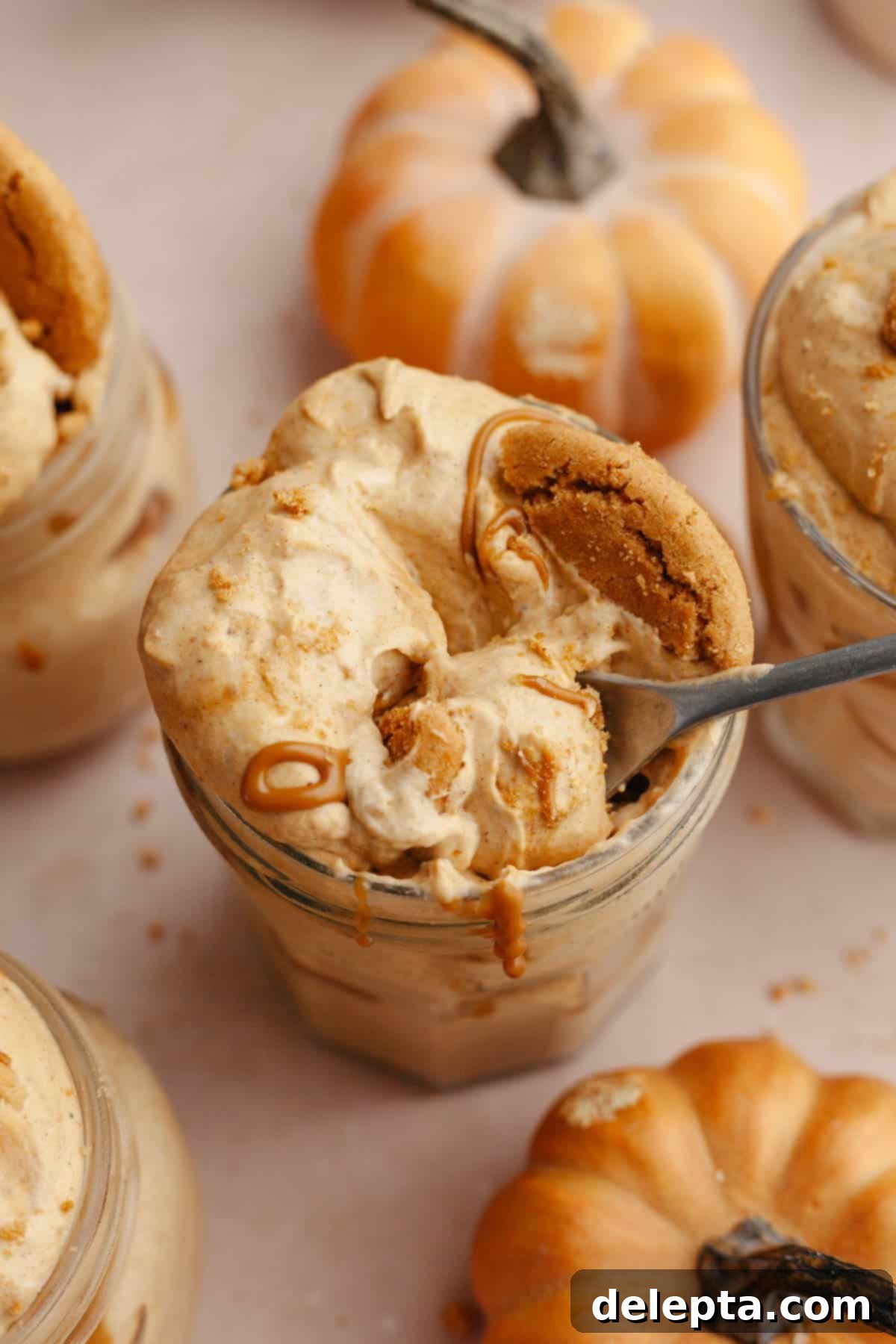 Effortless No Bake Pumpkin Delights 11 Two pumpkin pie cups in mason jars, topped with crushed gingersnap cookies and drizzled cookie butter. One jar has a spoon inserted, ready to be enjoyed.