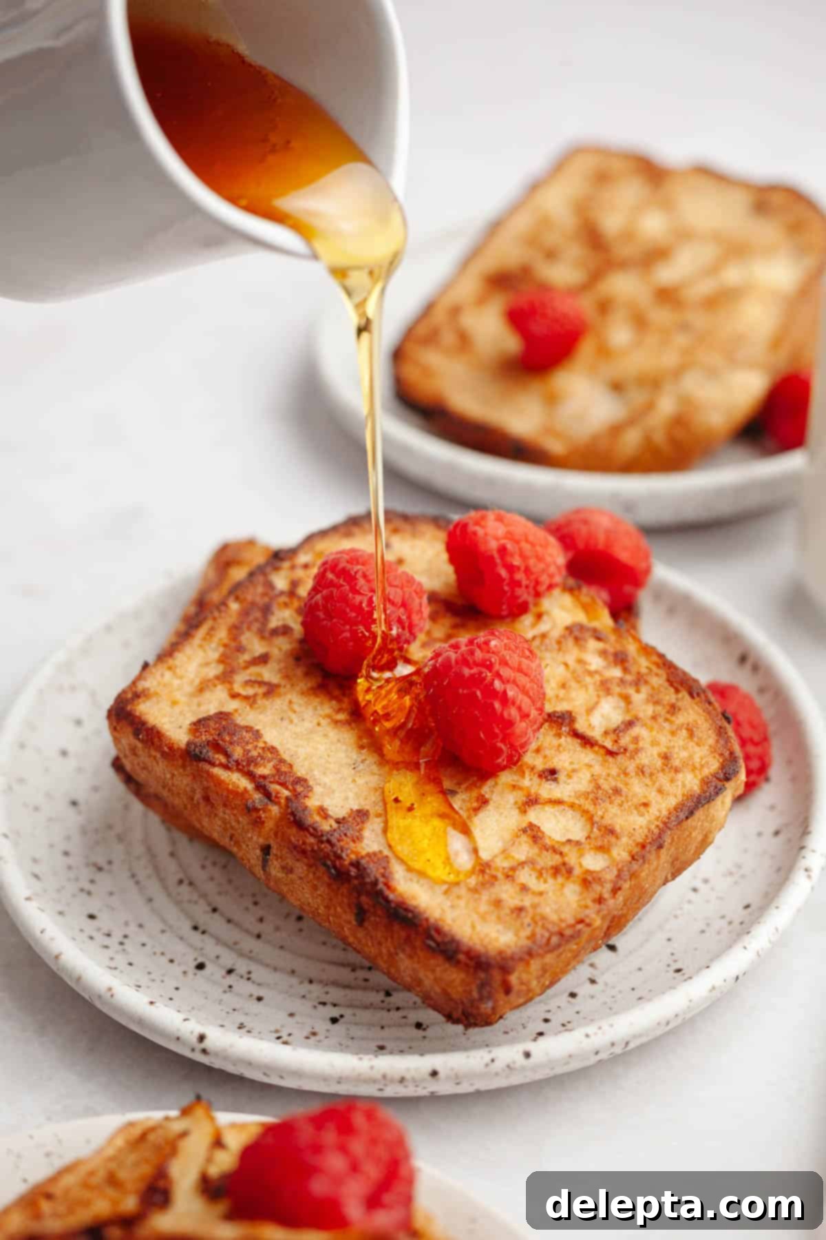 Effortless Buttermilk Brioche French Toast 2 Two slices of homemade buttermilk french toast on a small plate, topped with fresh raspberries and maple syrup being poured gracefully on top.
