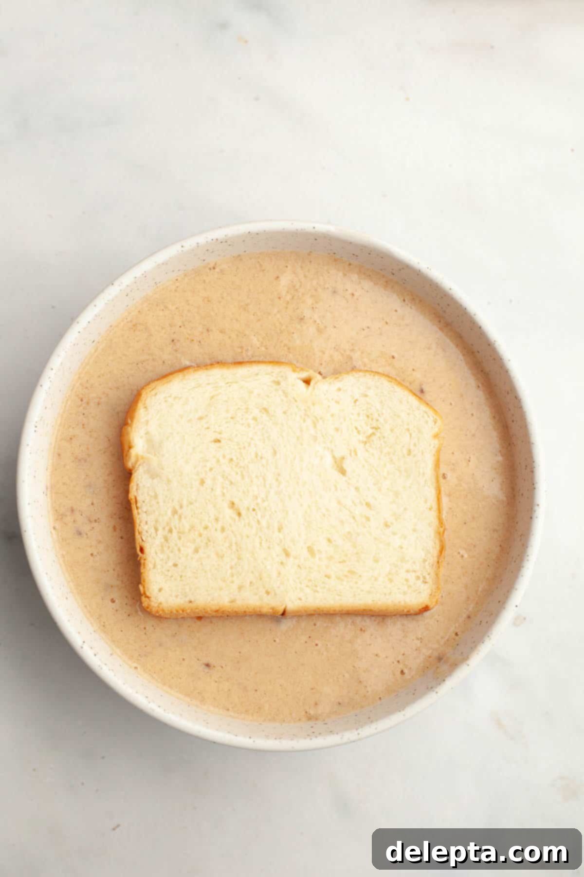 Effortless Buttermilk Brioche French Toast 5 A thick slice of brioche bread soaking in a bowl of creamy buttermilk french toast mixture.