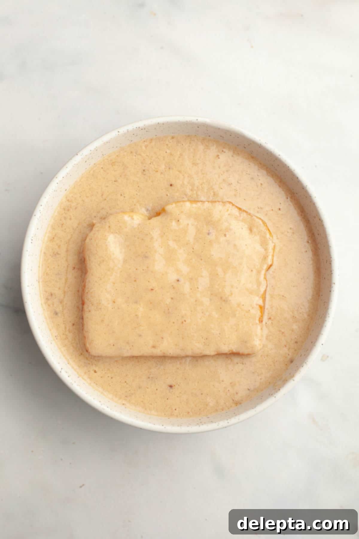 Effortless Buttermilk Brioche French Toast 6 A slice of brioche bread being flipped in a bowl to soak the other side in the rich French toast batter.