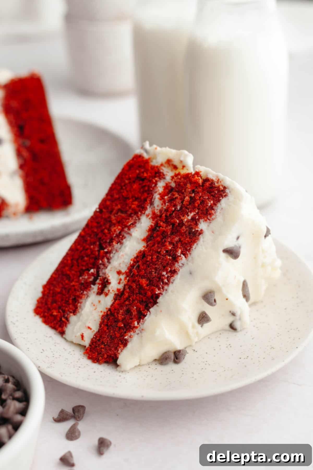 Red Velvet Chocolate Chip Indulgence 2 A perfectly sliced piece of red velvet chocolate chip cake, revealing its vibrant red layers and creamy frosting, resting on a small white plate.