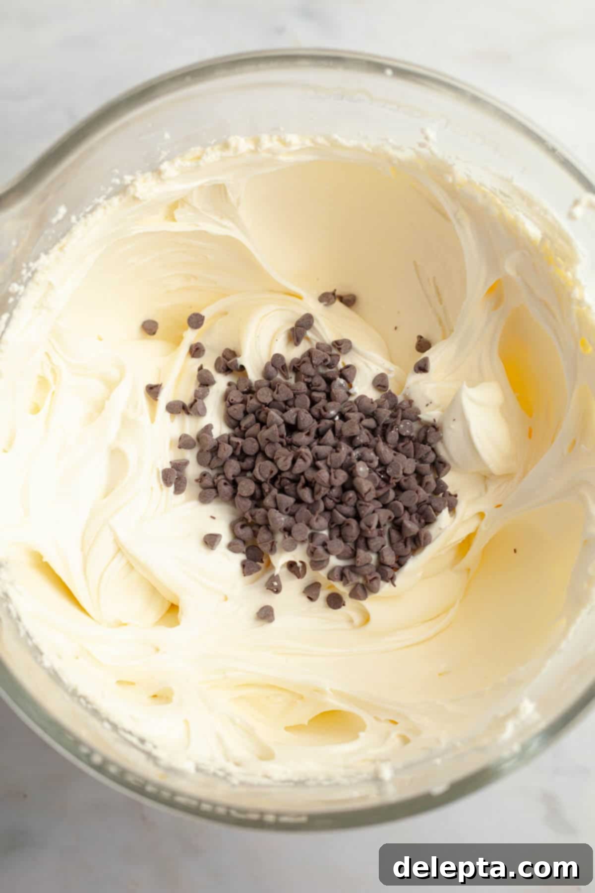 Red Velvet Chocolate Chip Indulgence 11 A large glass mixing bowl filled with creamy white cream cheese frosting, generously mixed with numerous mini chocolate chips, ready for cake assembly.