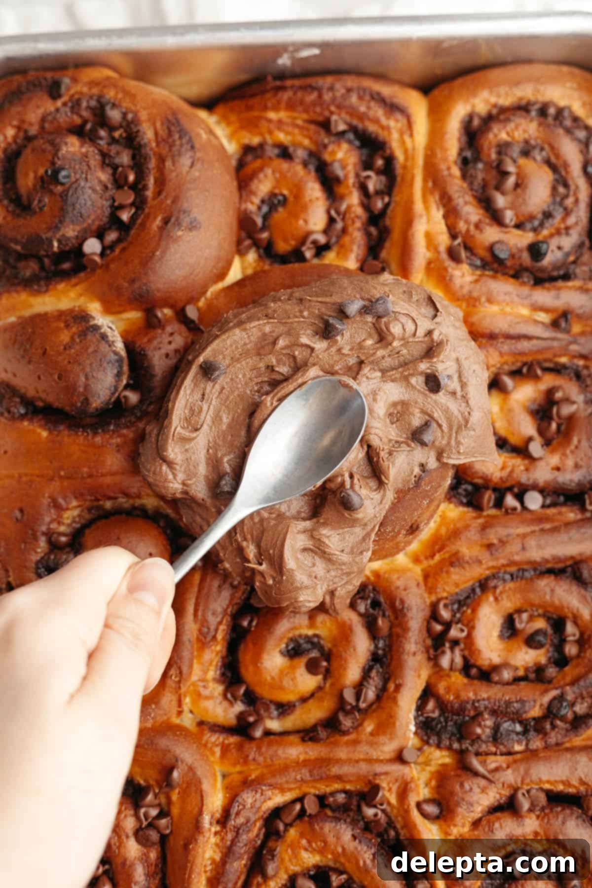 Decadent Double Chocolate Cinnamon Swirls 2 A hand is gently spreading thick, dark chocolate cream cheese frosting onto warm, freshly baked chocolate cinnamon rolls in a white ceramic baking dish.
