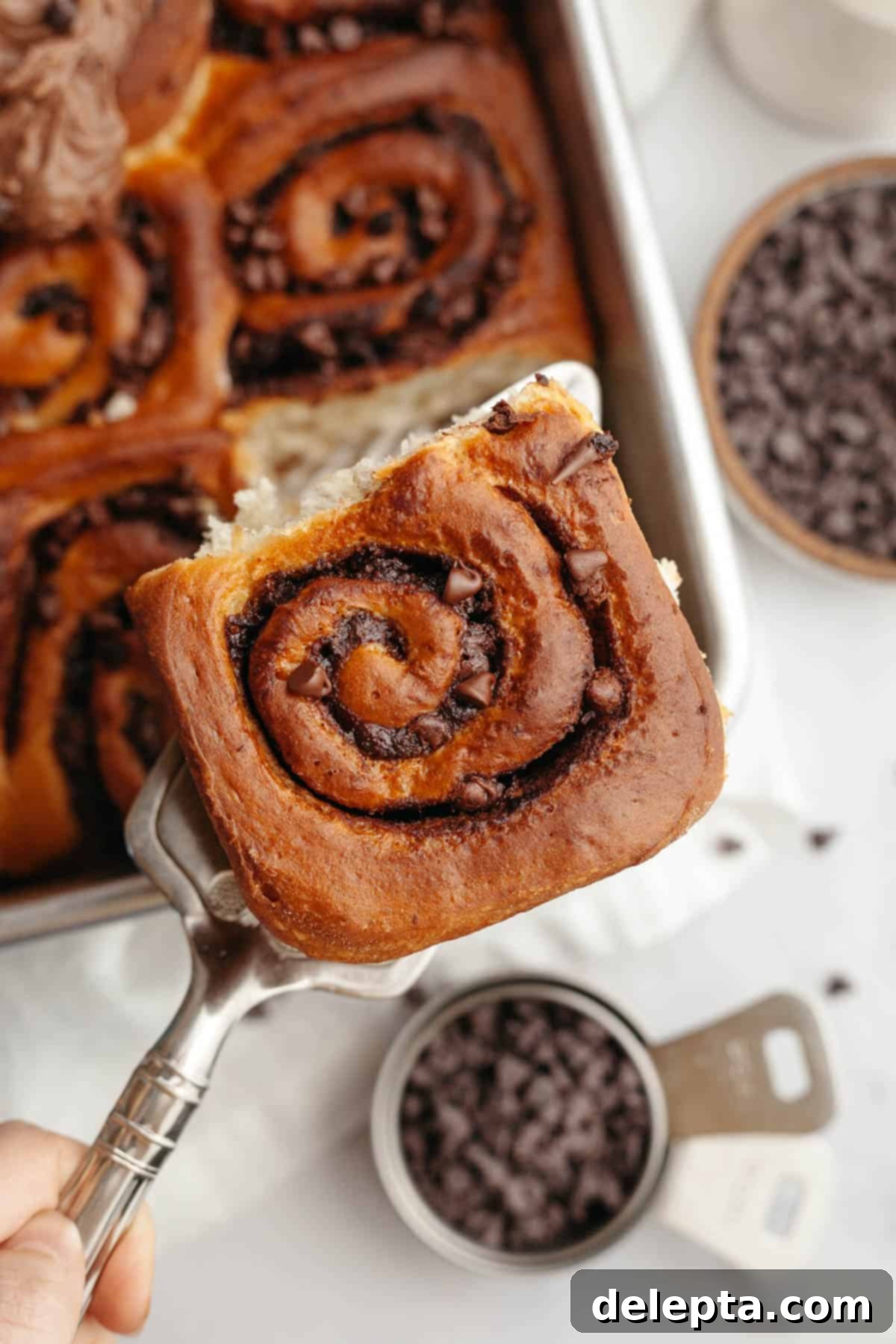 Decadent Double Chocolate Cinnamon Swirls 13 A silver cake server is carefully lifting a single gooey chocolate cinnamon roll from a baking pan, showing off its rich filling and frosting.