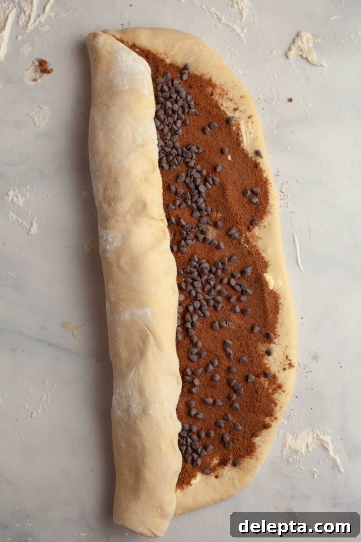 Decadent Double Chocolate Cinnamon Swirls 8 A person carefully rolling up a large rectangular sheet of dough filled with chocolate and cinnamon, forming a tight log.
