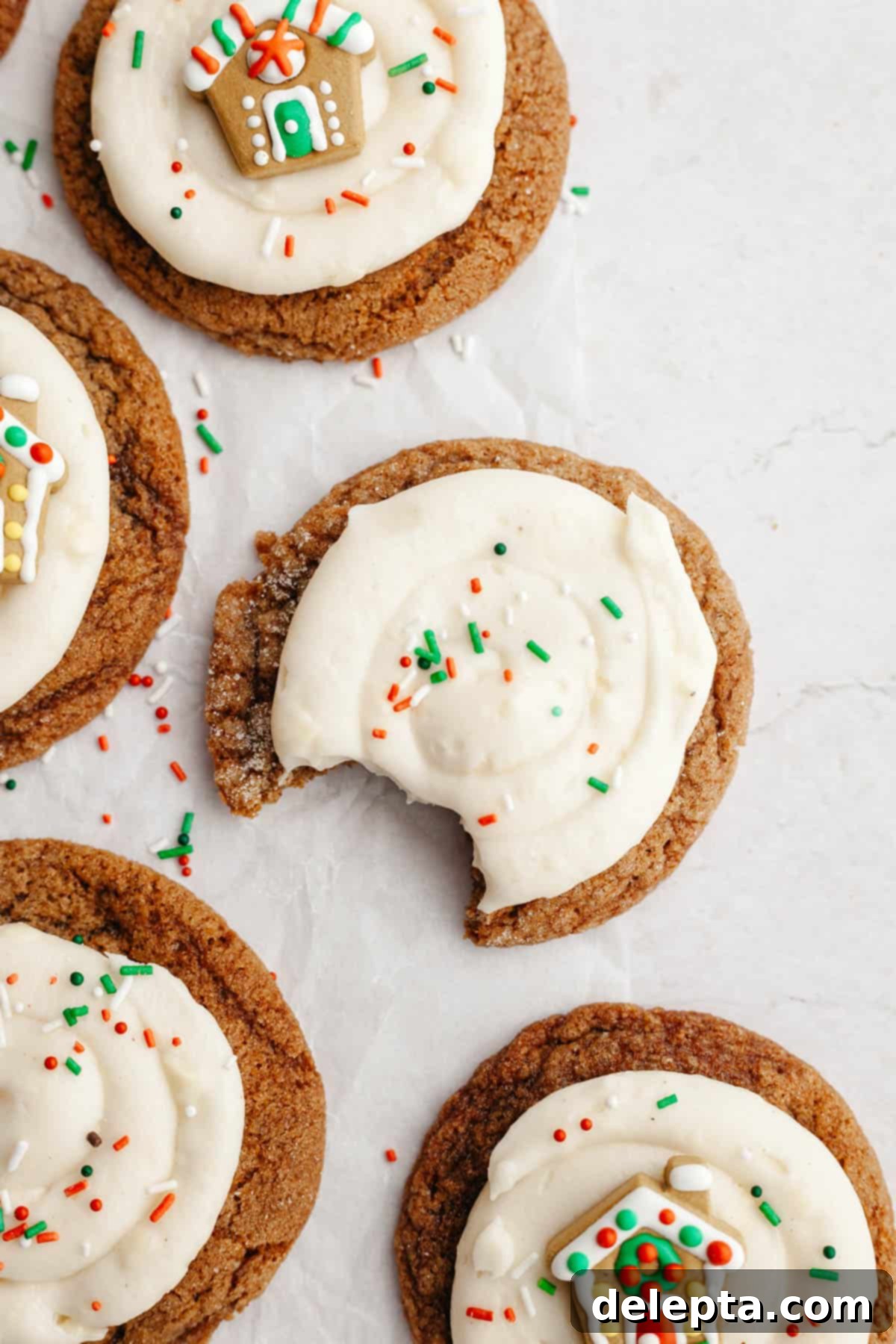 Velvet Gingerbread Cookies with Cream Cheese Frosting 11 A close-up of a frosted gingerbread cookie adorned with cream cheese frosting, a small gingerbread house decoration, and festive sprinkles.