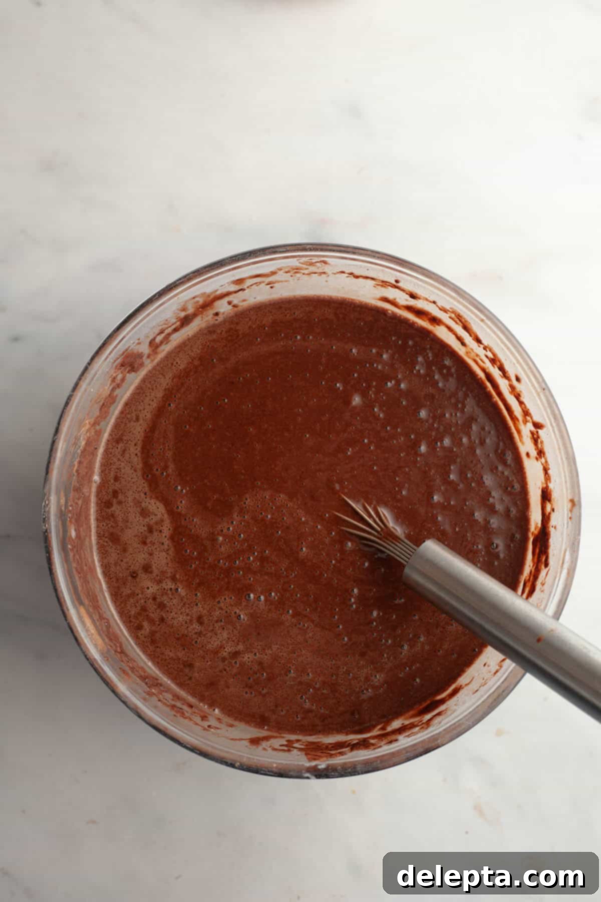 Moist and Easy Chocolate Cake with Decadent Chocolate Ganache 6 Smooth, dark chocolate cake batter filling a large glass bowl, with a silver whisk resting within, indicating it's just been mixed.