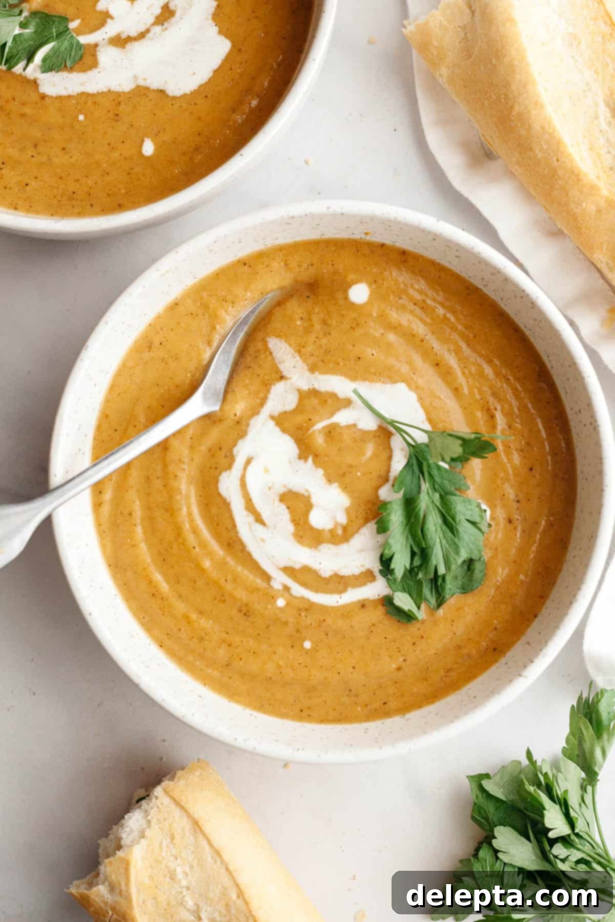 Velvety Roasted Butternut Cauliflower Soup 8 A white soup bowl with creamy butternut squash cauliflower soup, garnished with a fresh herb sprig and a swirl of cream, with a spoon in the bowl.
