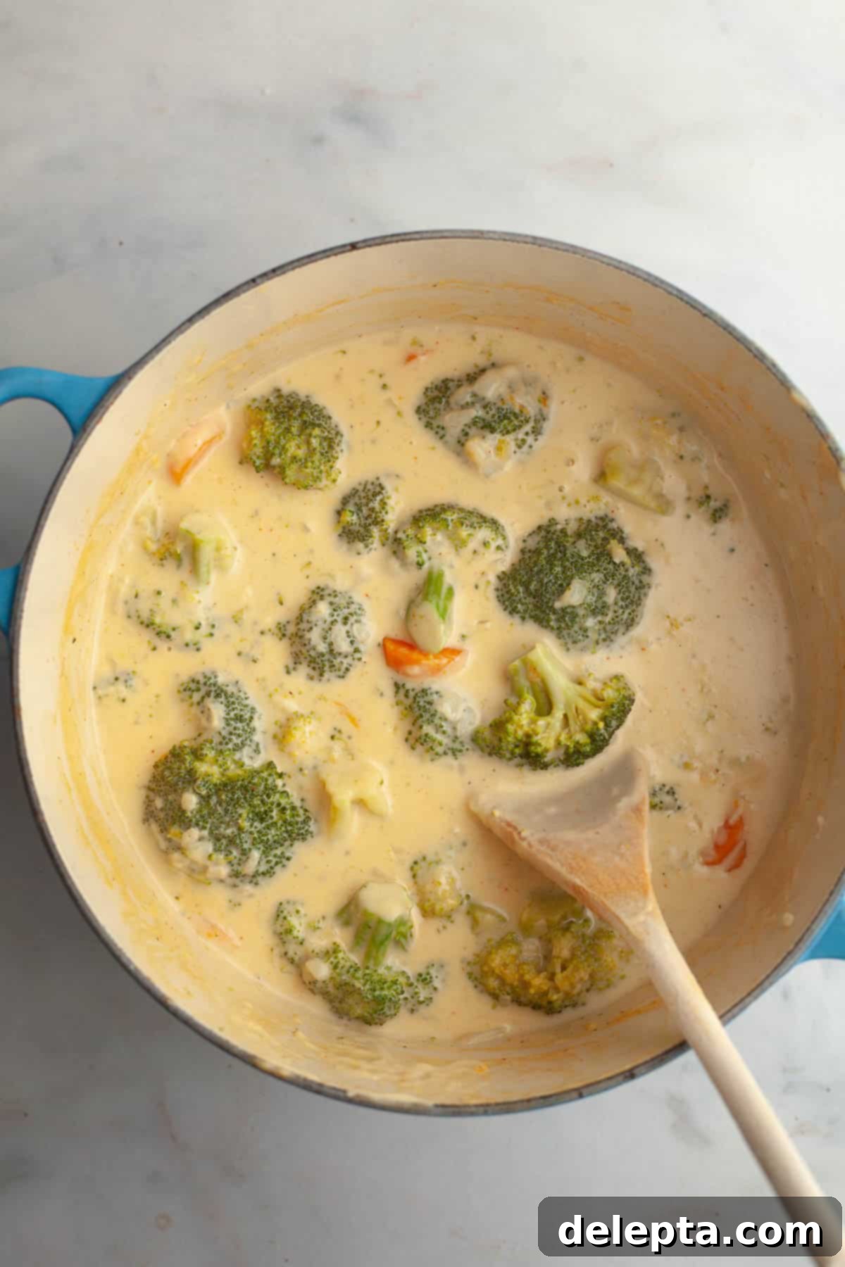 Velvety Chicken and Broccoli Cheddar Soup 7 A large dutch oven showing broccoli florets and chopped carrots simmering in the creamy soup base, with a wooden spoon resting in the pot.