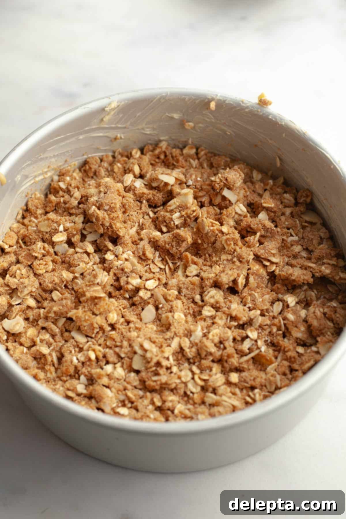 Irresistible Brown Butter Cardamom Almond Cake Recipe 9 A round cake pan with an unbaked cardamom almond cake covered in an oaty streusel.