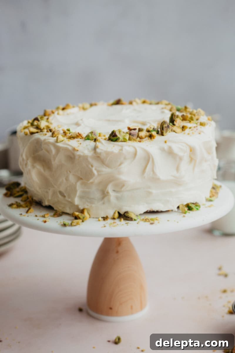 Pistachio-Infused Carrot Cake with Cloud-Like Cream Cheese Frosting 7 A frosted cake decorated with pistachios on a white and wooden cake stand.