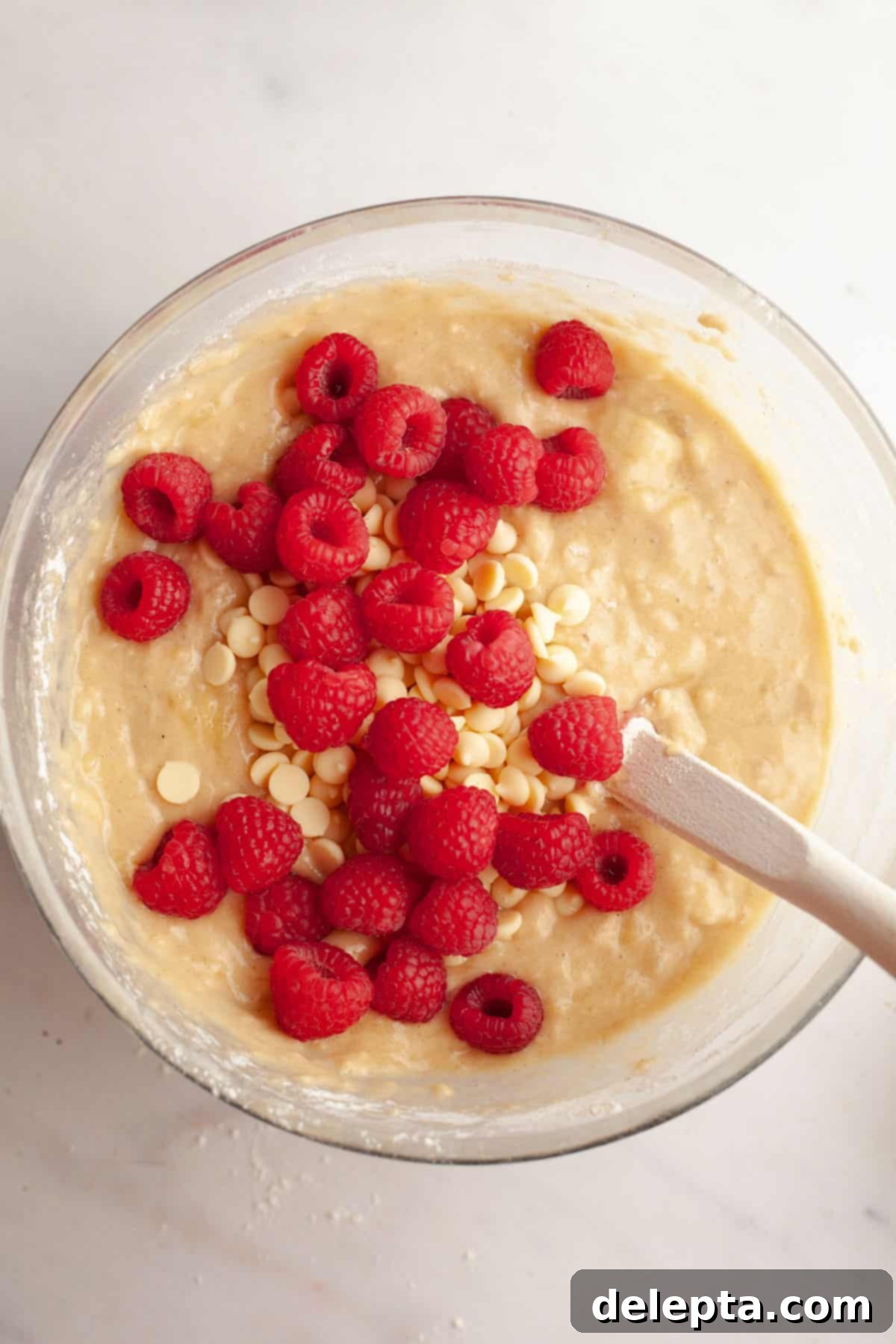 Effortless Banana Raspberry White Chocolate Chip Muffins 7 A large glass bowl with muffin batter in it, topped with fresh raspberries and white chocolate chips, ready to be folded in.