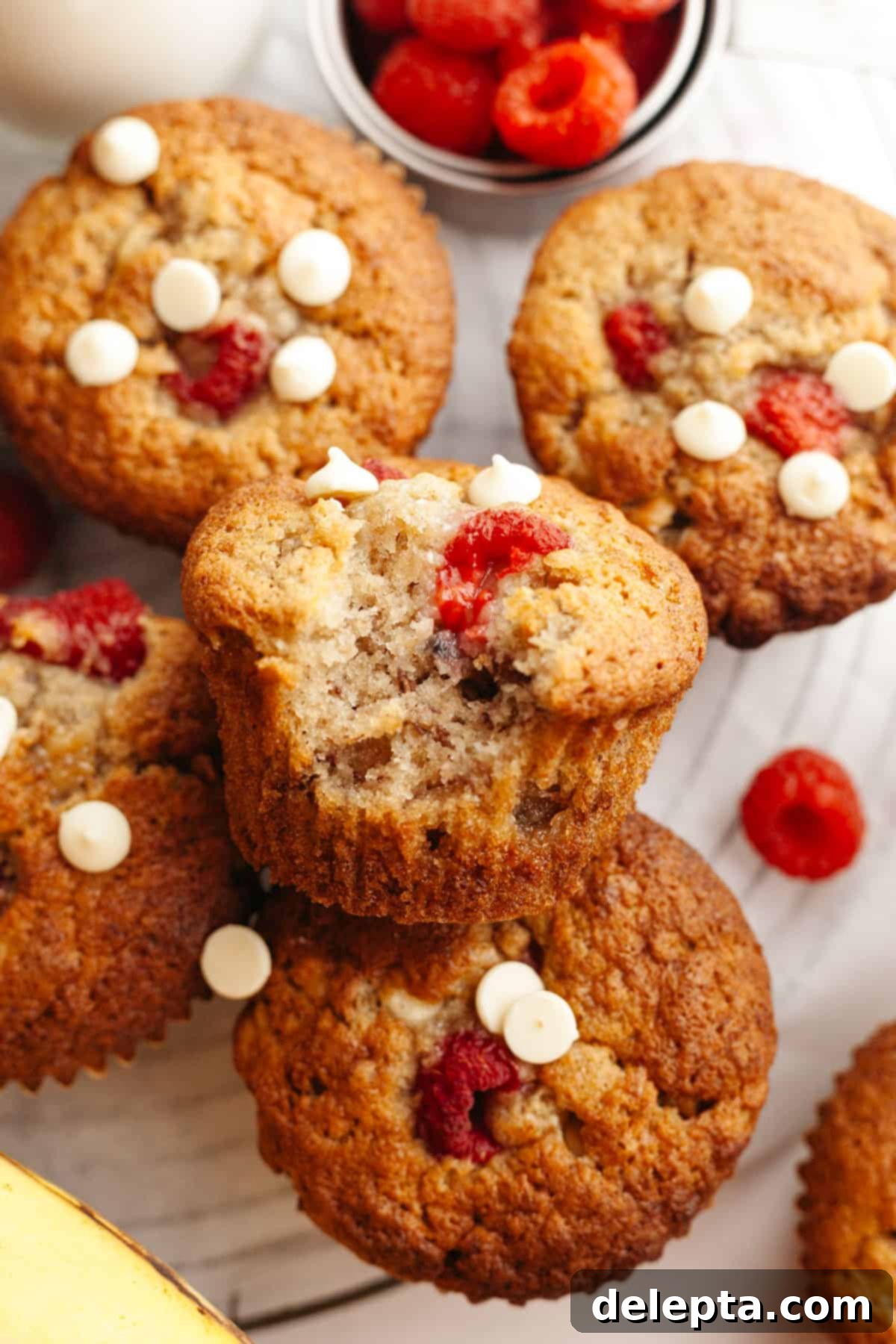 Effortless Banana Raspberry White Chocolate Chip Muffins 10 A close-up view of freshly baked banana raspberry muffins with glistening white chocolate chips, one with a delectable bite removed, revealing the soft interior.