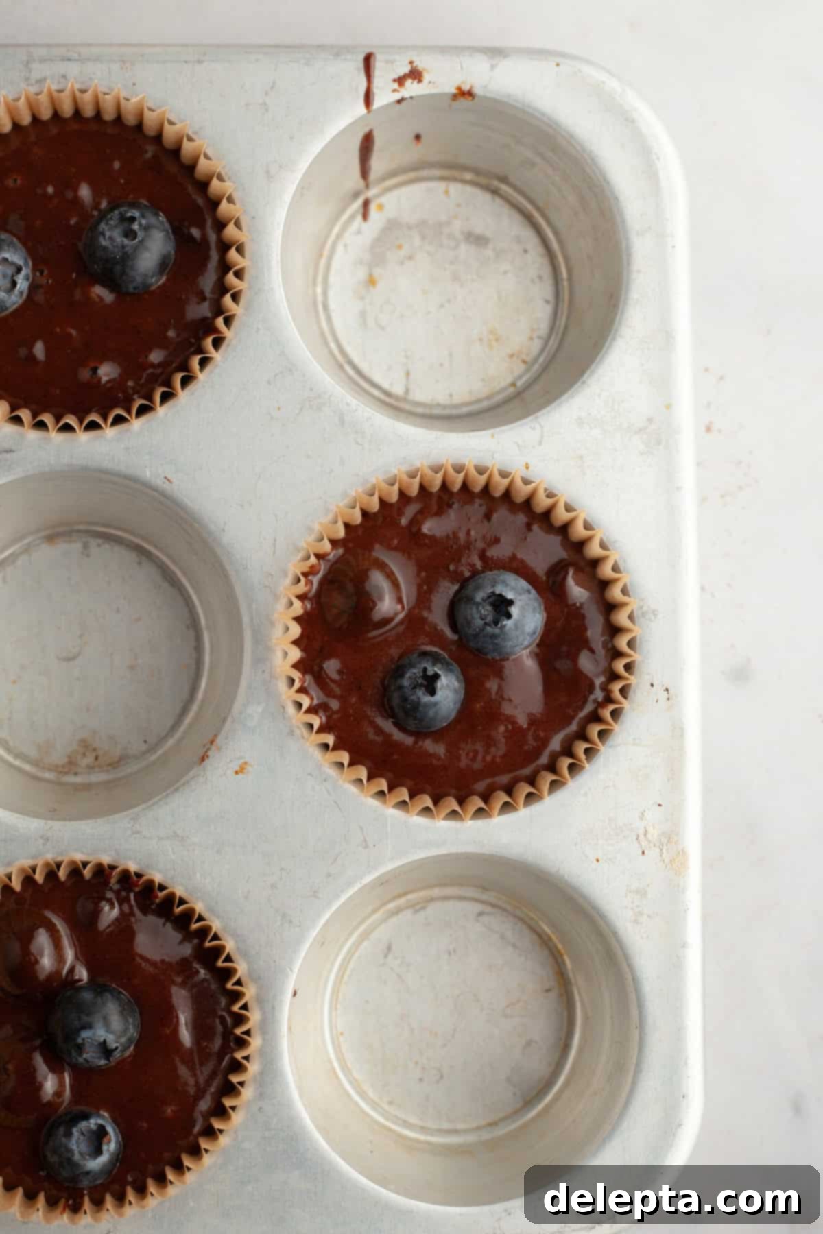 Decadent Blueberry Chocolate Sour Cream Muffins 7 A muffin pan filled with unbaked chocolate blueberry muffin batter in paper liners, spaced out to promote better rising and a bakery-style dome.