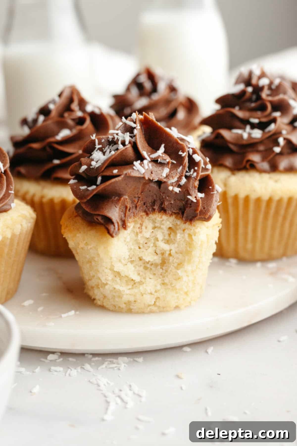 Double Chocolate Coconut Indulgence 2 Coconut cupcakes with chocolate frosting, sprinkled with shredded coconut. One cupcake has a bite taken out of it.