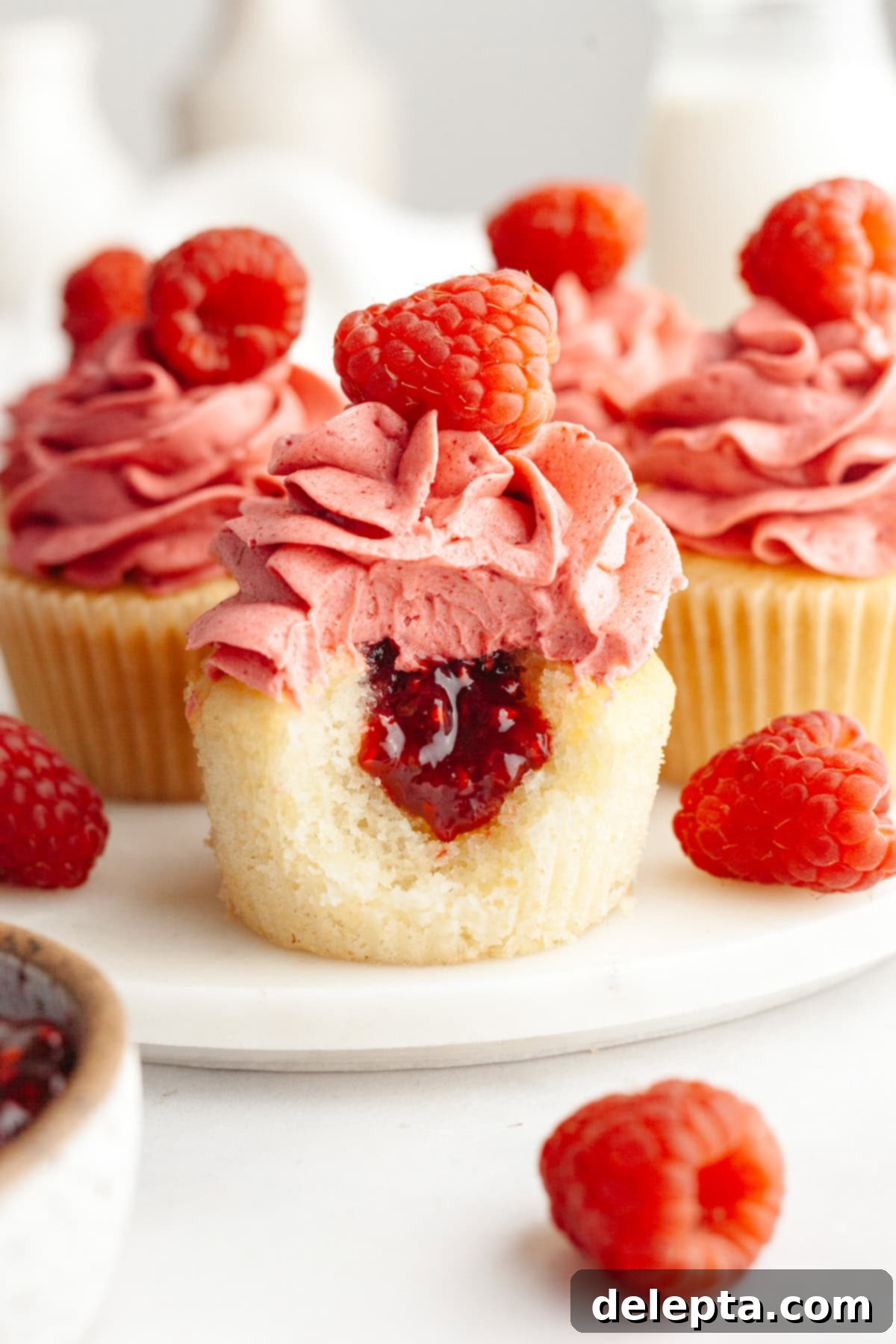 Raspberry Almond Jam Burst Cupcakes 2 A close up of an almond cupcake frosted with raspberry buttercream, it has a bite taken out of it showing a jam center. The cupcake is sitting on a white plate with other cupcakes blurred in the background.