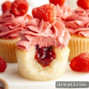 Raspberry Almond Jam Burst Cupcakes 11 A close up of an almond cupcake frosted with raspberry buttercream, it has a bite taken out of it showing a jam center.