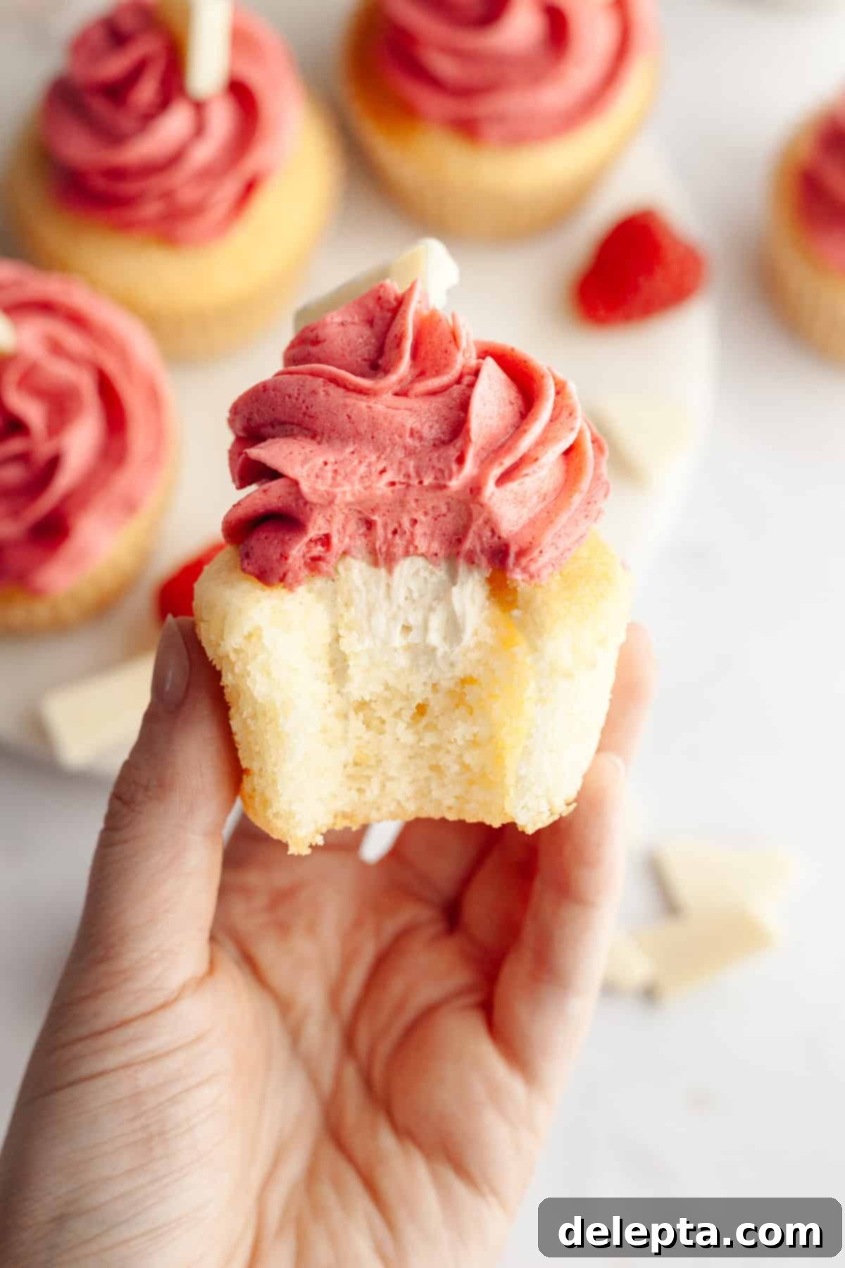 Decadent Raspberry White Chocolate Ganache Cupcakes 13 A hand holding a vanilla cupcake with a bite taken out of it, it has a white chocolate ganache filling and pink frosting.