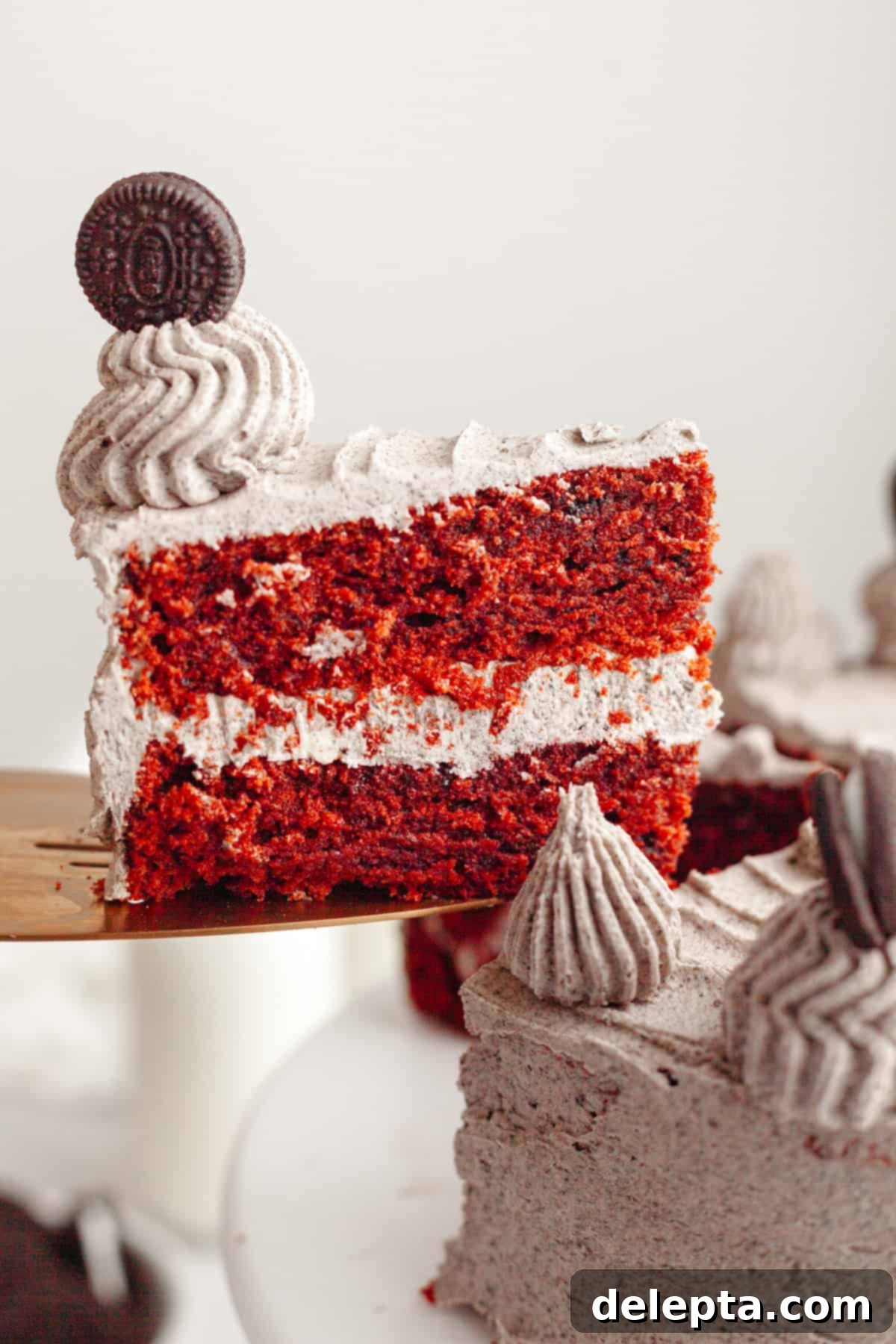Red Velvet Oreo Cake with Oreo Buttercream 2 A slice of Red Velvet Oreo Cake being lifted out with a cake server, showcasing the vibrant red layers and speckled Oreo frosting.
