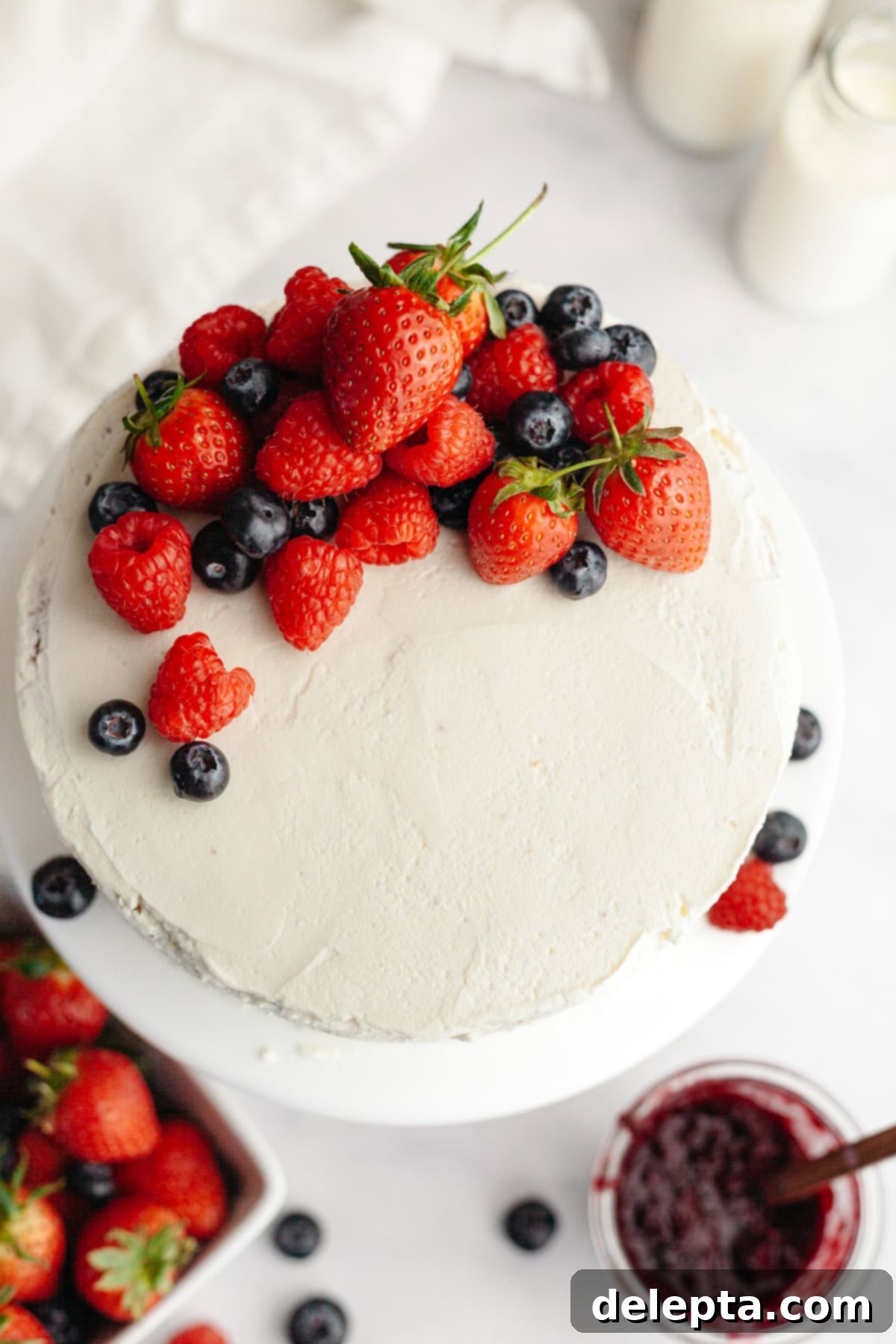 Berry Vanilla Mascarpone Bliss 12 A magnificent round berry vanilla cake, beautifully frosted in white and adorned with fresh strawberries, raspberries, and blueberries. It rests on a pristine white surface, accompanied by a bowl of assorted berries and a charming jar of jam, highlighting its fresh and fruity essence.