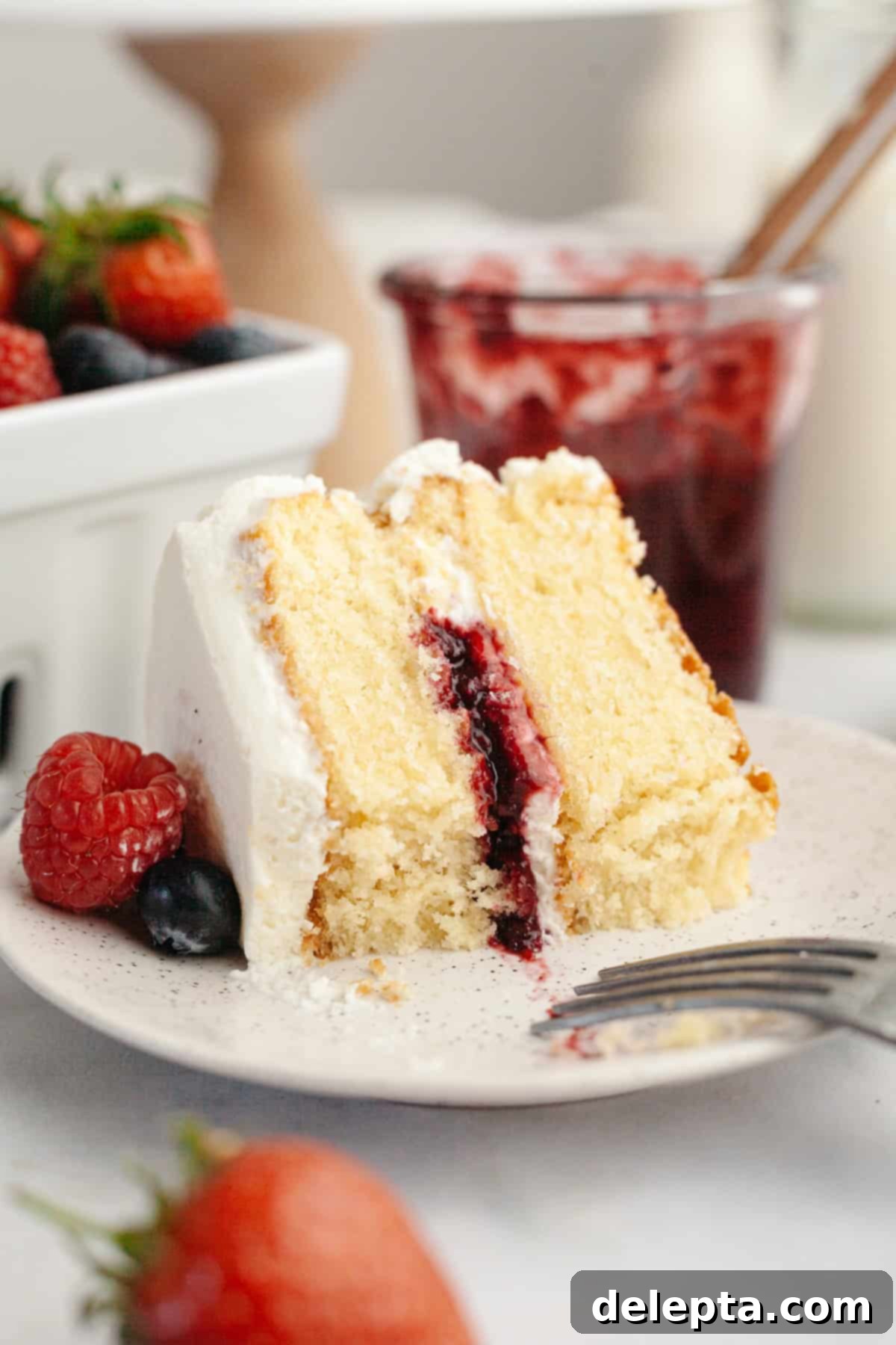 Berry Vanilla Mascarpone Bliss 13 A perfect slice of berry vanilla layer cake with white mascarpone frosting, a vibrant berry jam filling, and crowned with a fresh raspberry and blueberry. It sits elegantly on a plate, with additional strawberries and a jam jar artfully blurred in the background, inviting a delicious taste.