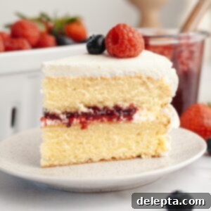 Berry Vanilla Mascarpone Bliss 14 A slice of a berry vanilla layer cake with white frosting, berry jam filling, and topped with a raspberry and blueberry, on a plate with strawberries and jam jar in the background.