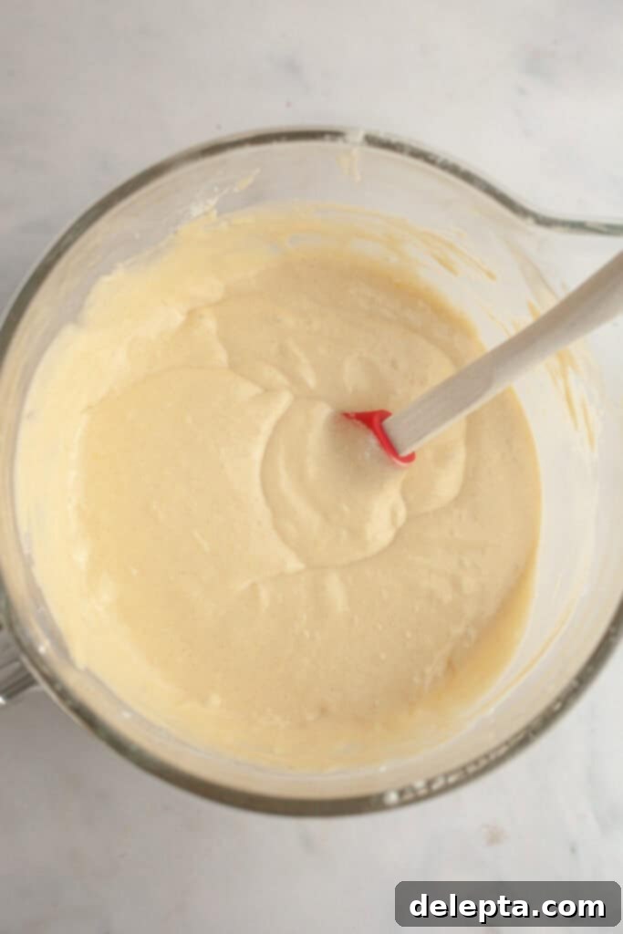 Berry Vanilla Mascarpone Bliss 5 Glass mixing bowl filled with smooth, pale vanilla cake batter, with a red and white spatula resting inside on a light countertop, indicating readiness for baking.