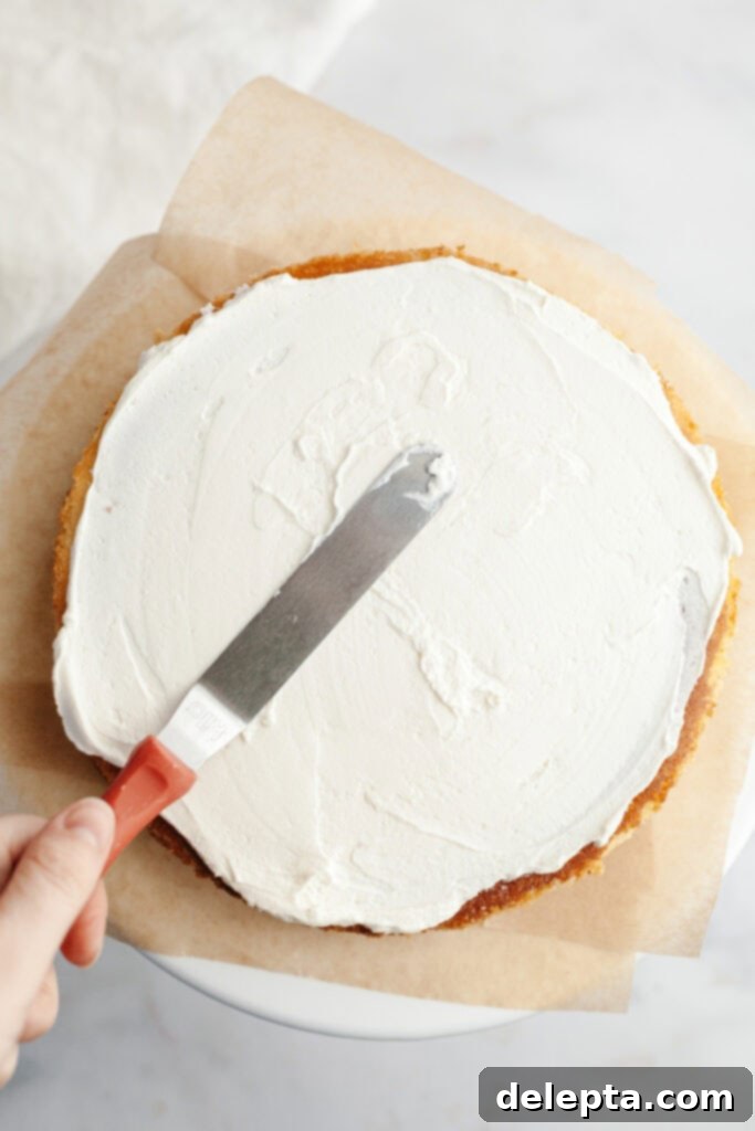 Berry Vanilla Mascarpone Bliss 9 A vanilla cake layer on parchment paper, with a hand spreading luscious mascarpone whipped cream frosting using a small offset spatula.