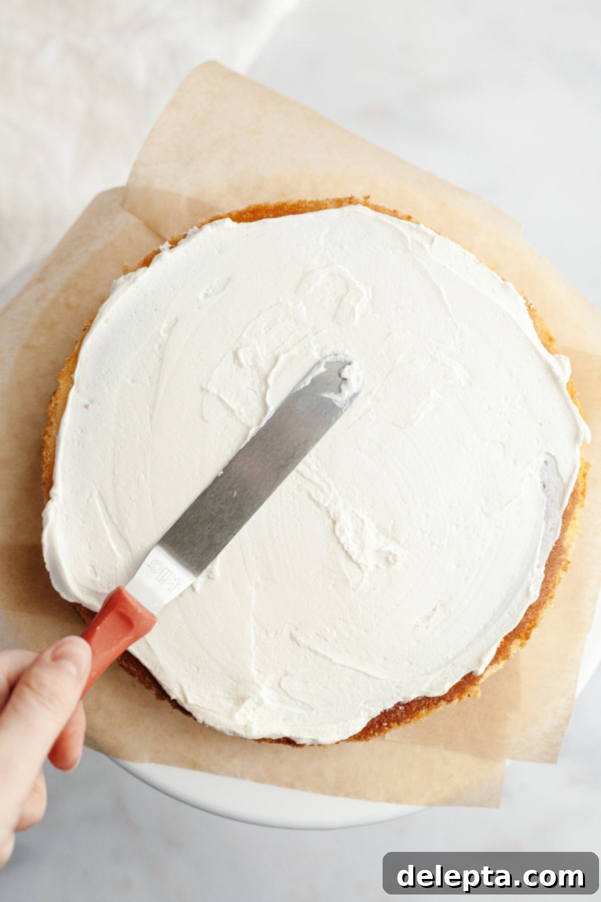 Dreamy 5-Ingredient Mascarpone Whipped Cream Frosting 9 A vanilla cake layer is on parchment paper, a hand is spread over whipped mascarpone frosting with a small offset spatula.