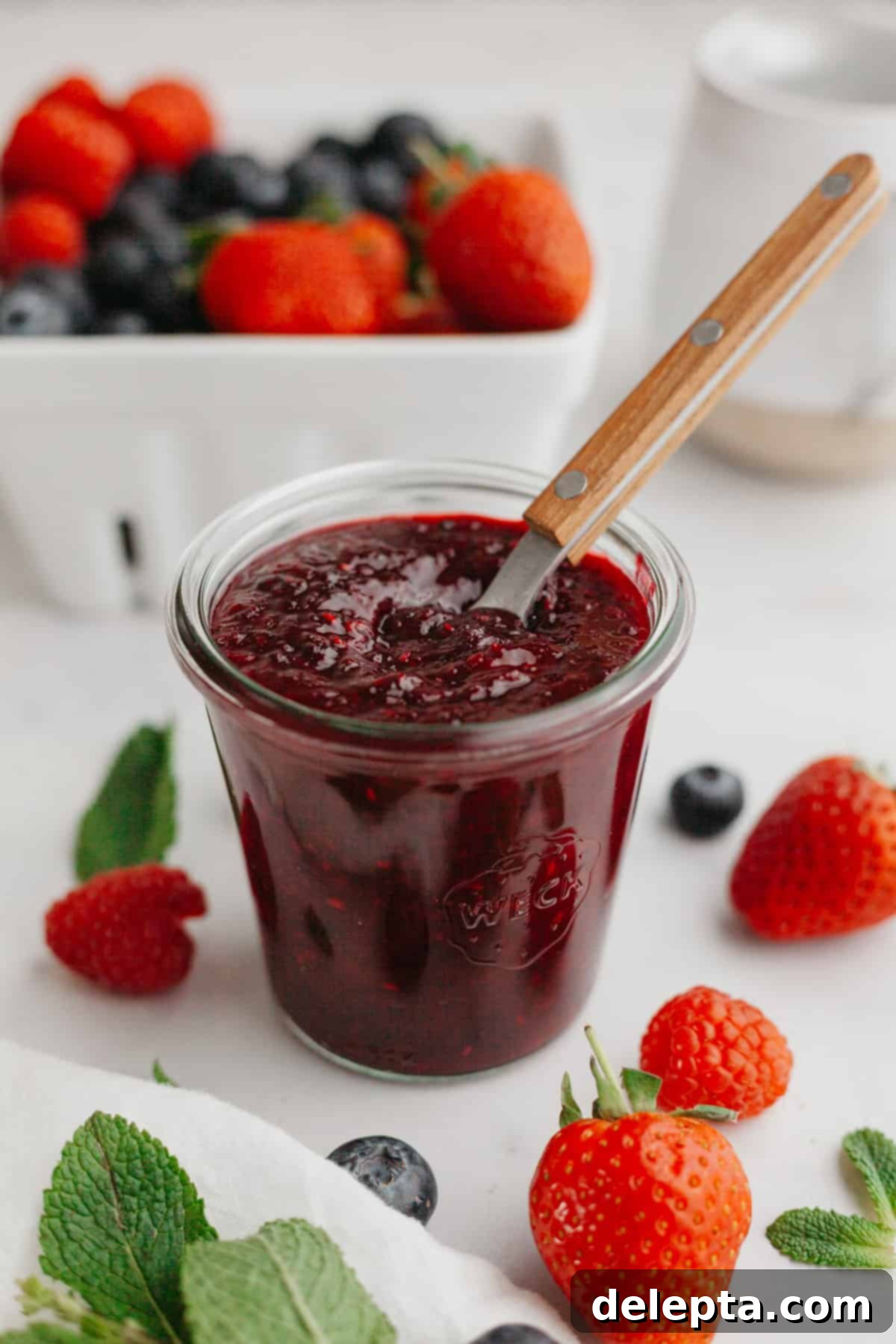 Quick Berry Compote with Frozen Fruit 2 Mixed berry compote in a glass jam jar with a wooden spoon. There are fresh berries and mint leaves in the background.