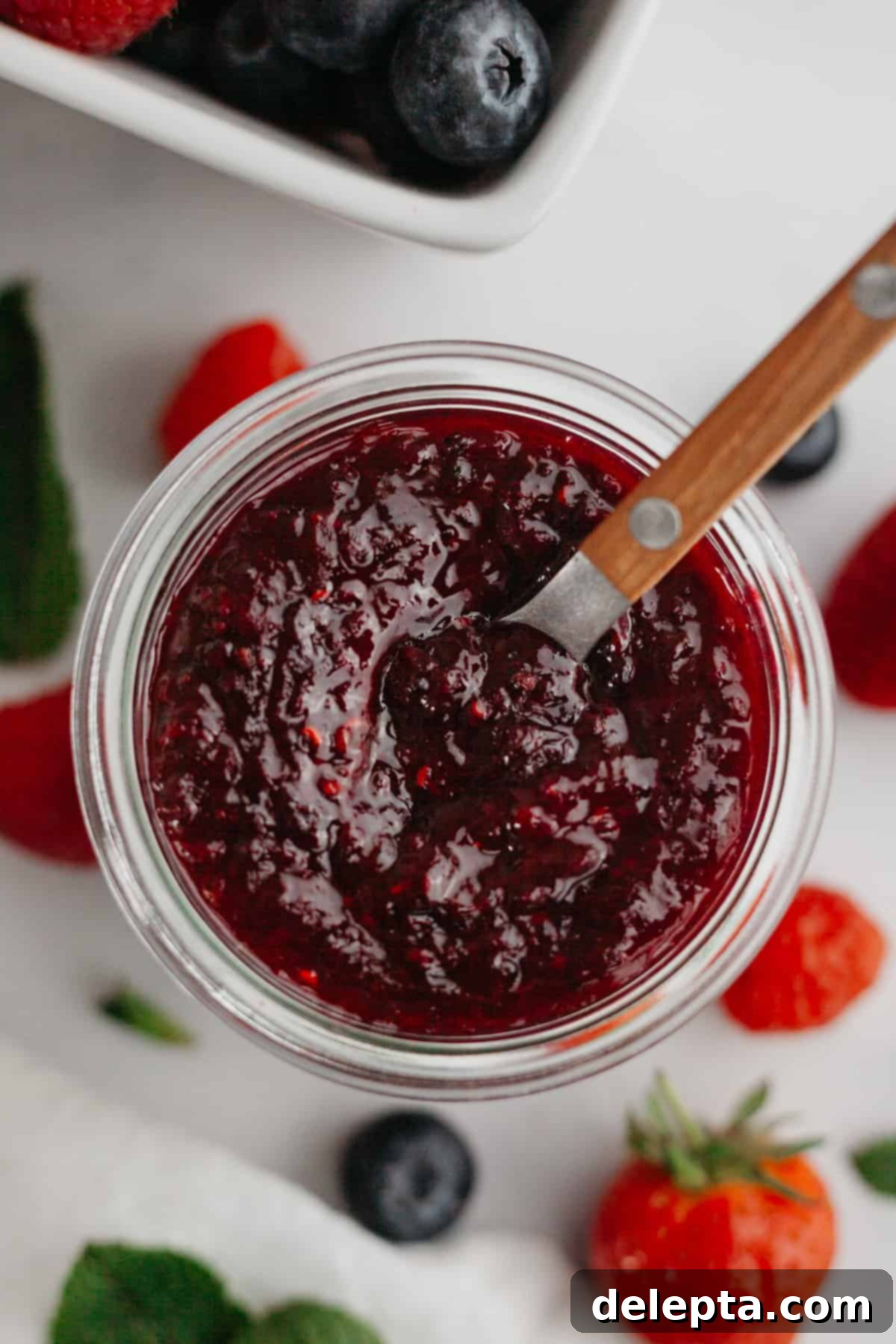Quick Berry Compote with Frozen Fruit 6 Mixed berry compote in a glass jam jar with a wooden spoon. There are fresh berries in the background.