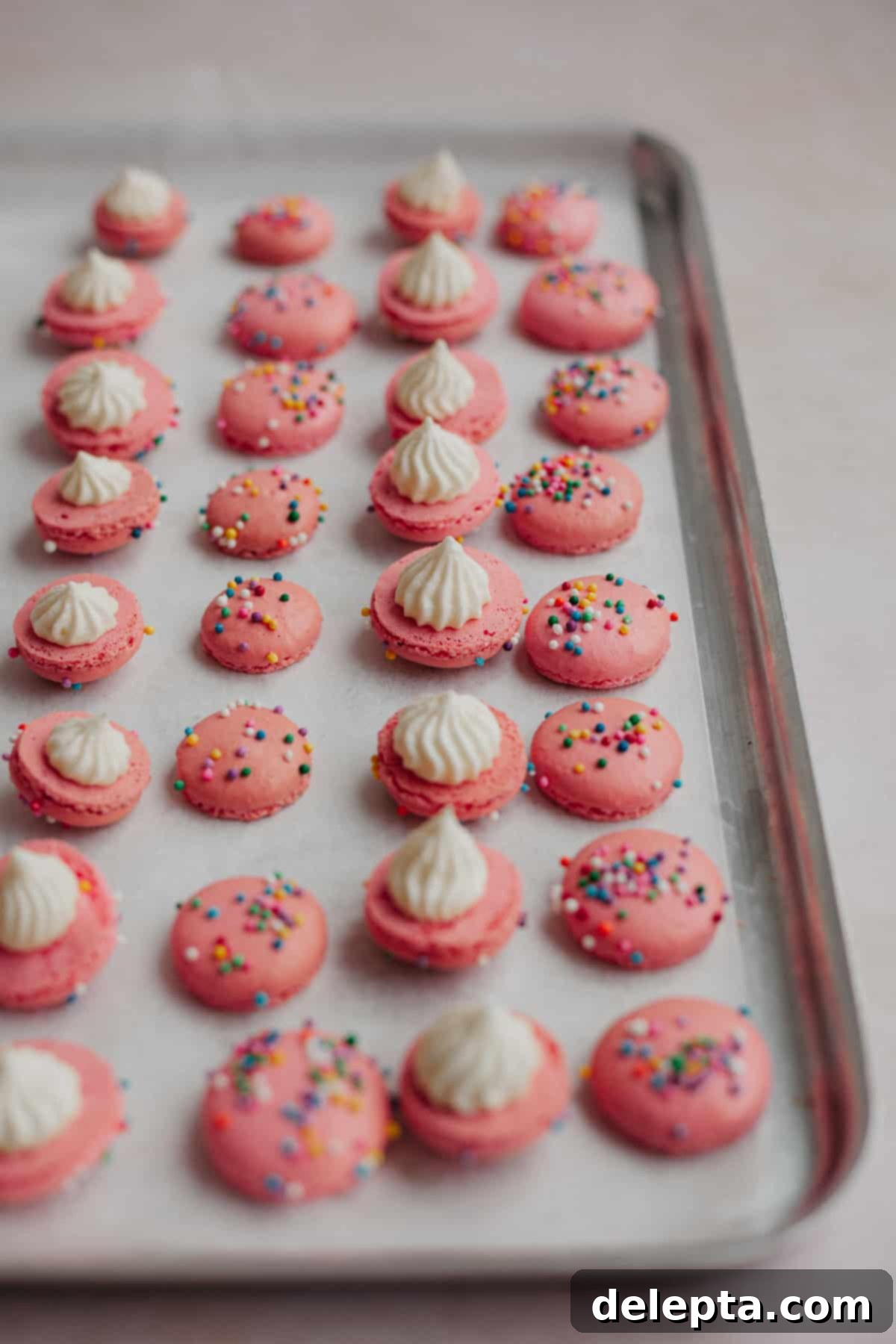 Funfetti Macarons 12 A baking tray lined with white parchment paper. It is filled with rows of macaron shells. Every other row, the shells are upside down and have a dollop of buttercream on them