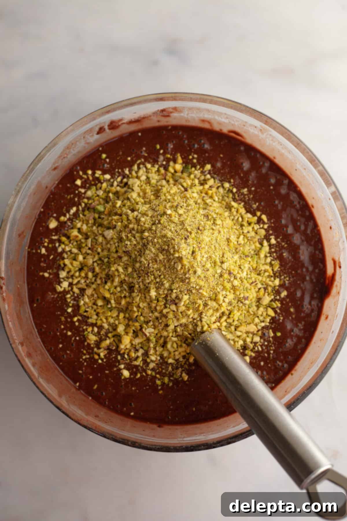 Decadent Chocolate Pistachio Dream Cake 6 Chocolate cake batter with crushed pistachios, being stirred with a silver whisk in a glass bowl.