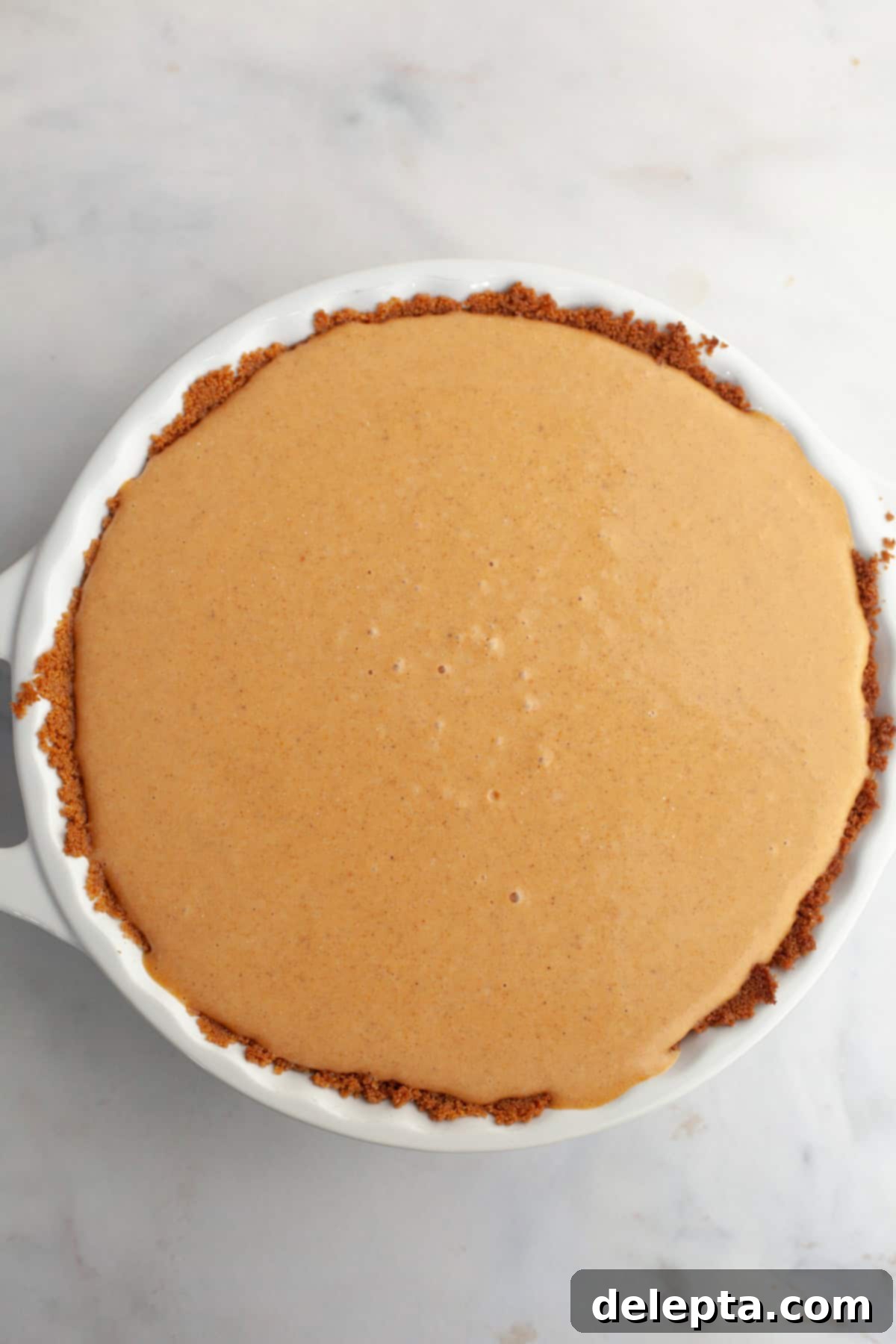 Decadent Brown Butter Pumpkin Pie with Gingersnap Perfection 8 An unbaked brown butter pumpkin pie in a white ceramic baking dish, ready for the oven, with its smooth surface and golden gingersnap crust.