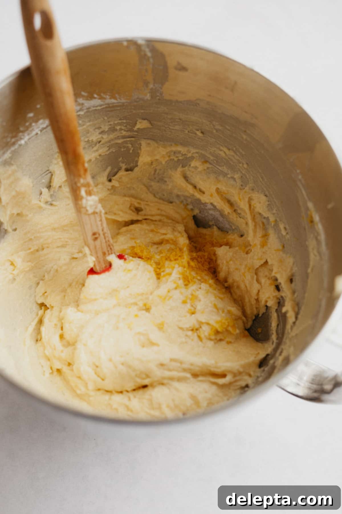 Sun-Kissed Blueberry Almond Lemon Cake 5 A large mixing bowl with smooth, creamy cake batter and a spatula with a wooden handle resting in it.